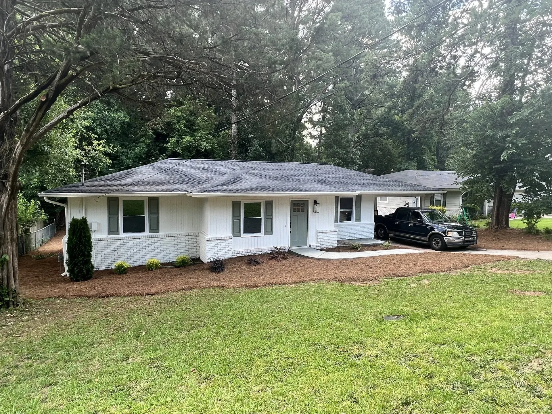 White house with a dark car parked in the driveway, surrounded by green grass and trees.