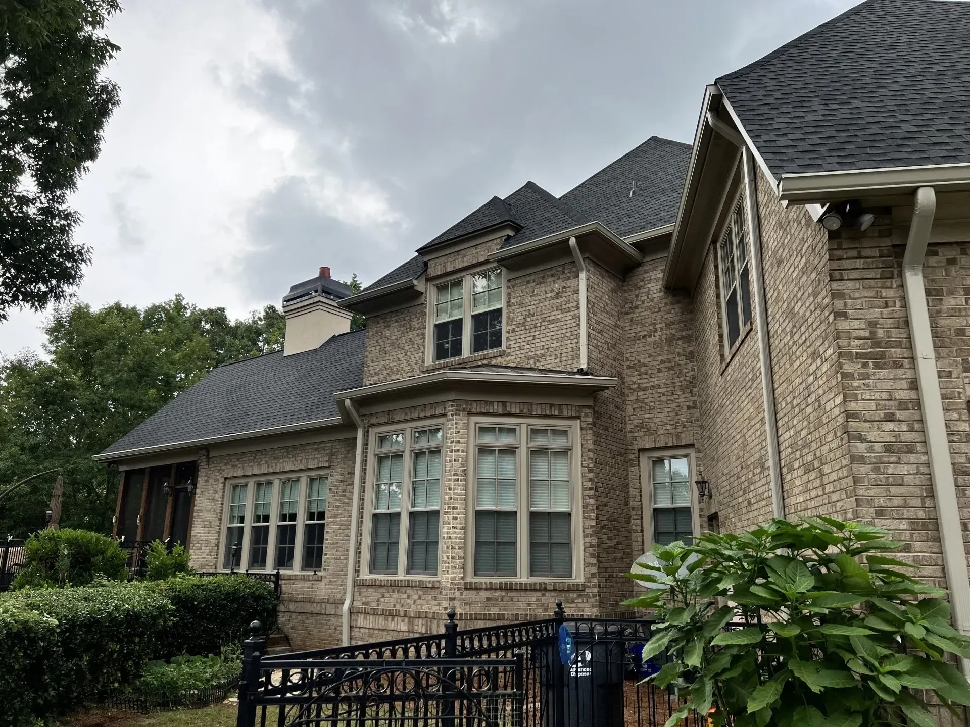 Brick house exterior with dark roof and windows under a cloudy sky.