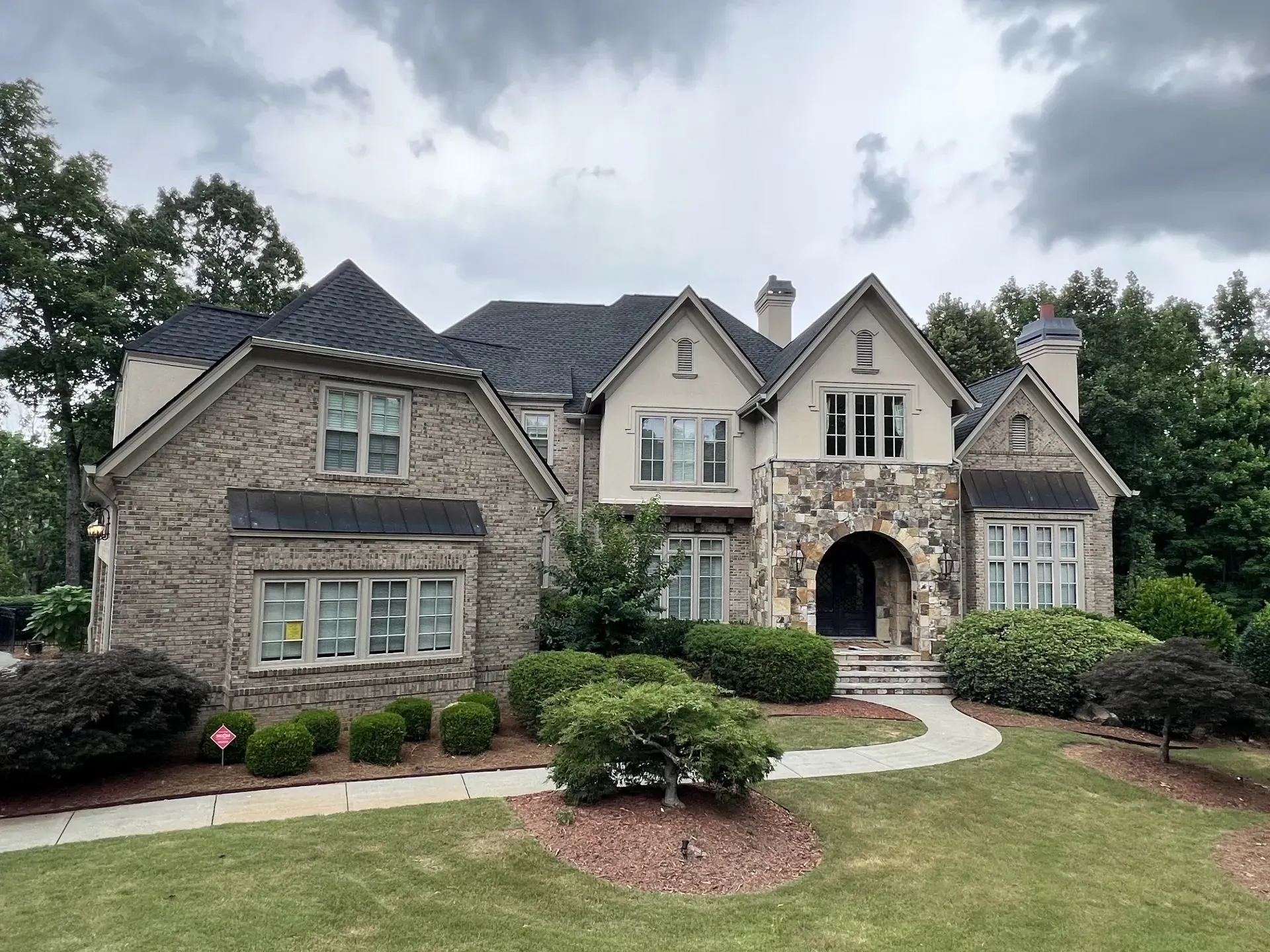 Two-story house with stone and brick exterior; arched entryway; green lawn and landscaping. Overcast sky.
