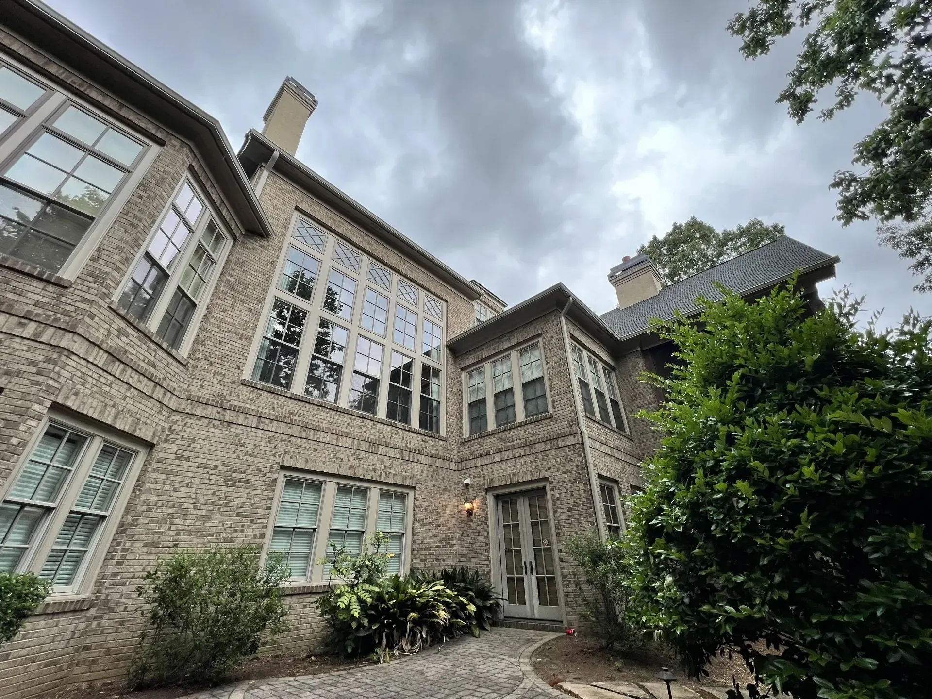 Brick house with large windows, under cloudy sky, surrounded by greenery.
