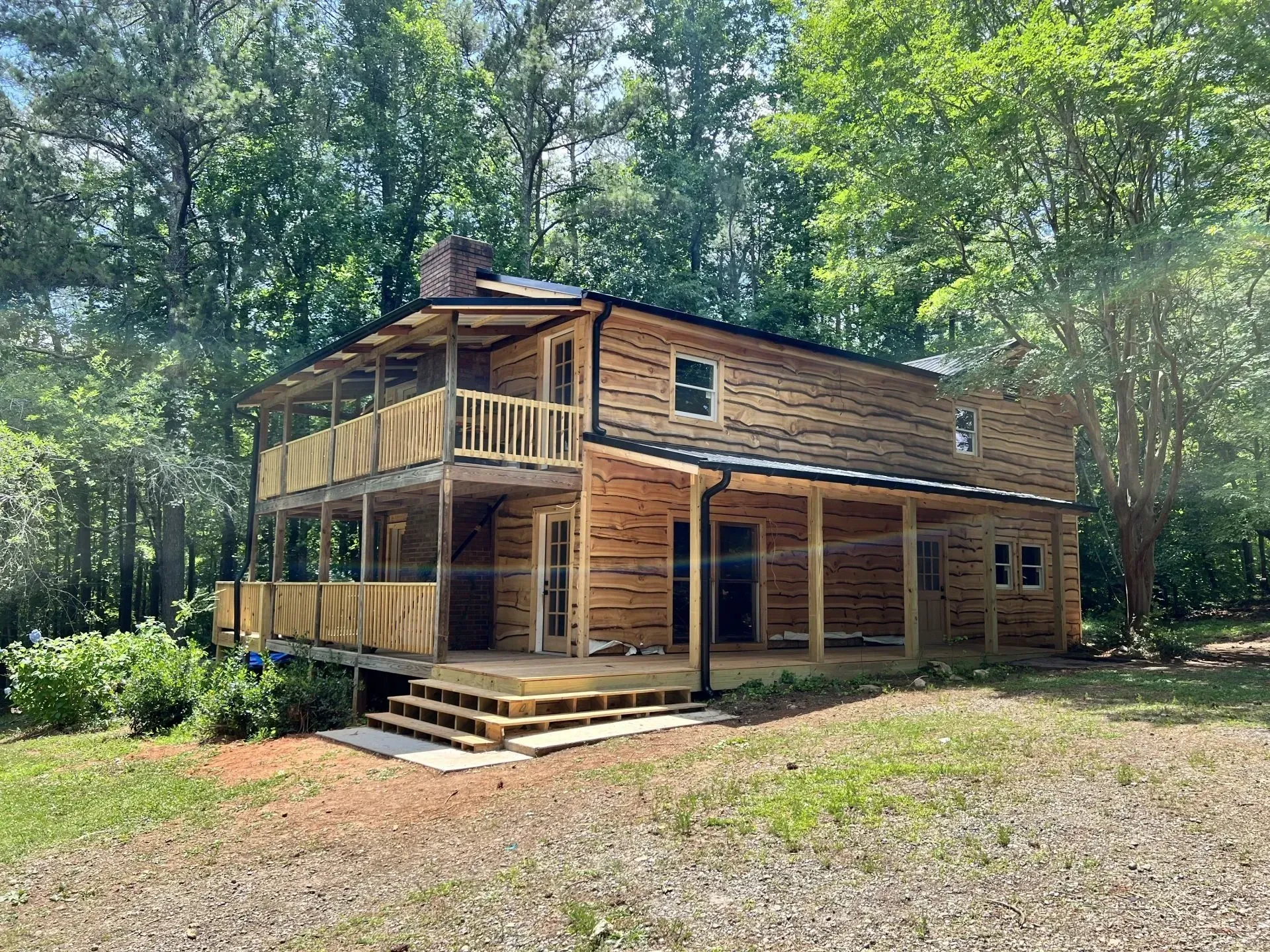 Two-story wooden house with wrap-around porch and forest backdrop.