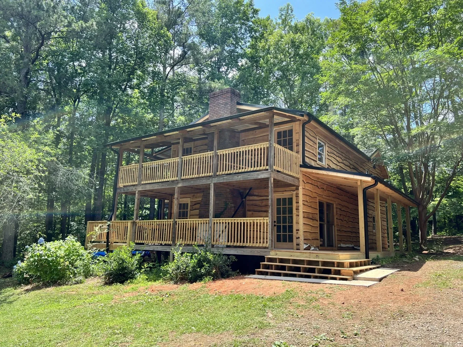Two-story log cabin with wooden balconies, set in a wooded area, with a grassy yard.