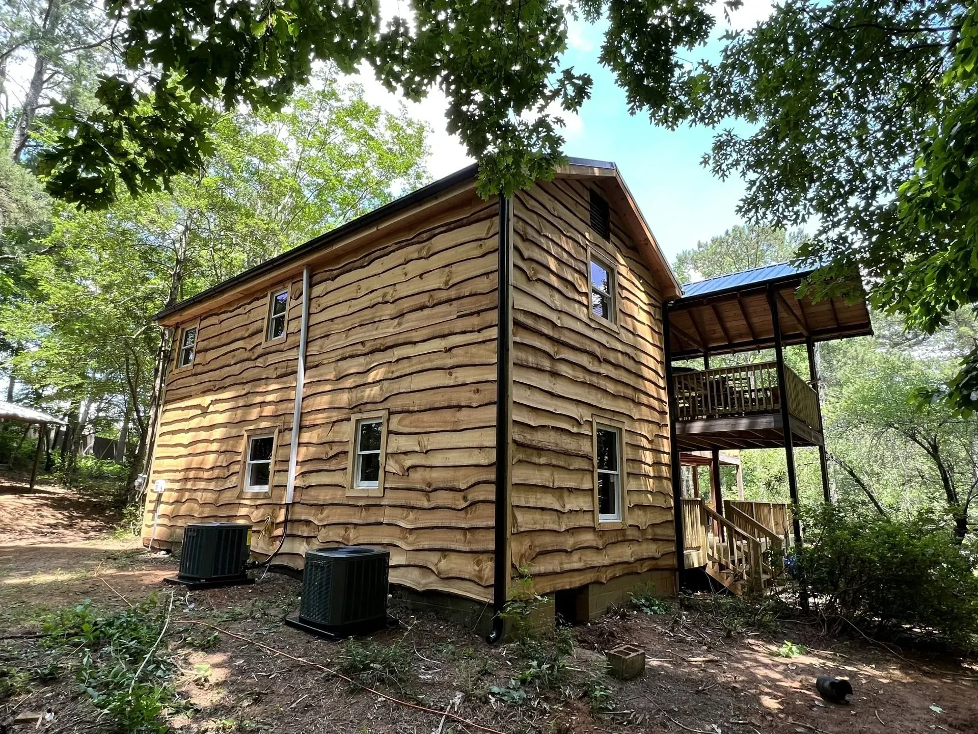 Two-story wooden house with a deck nestled amongst trees. Dark air conditioning units in front.