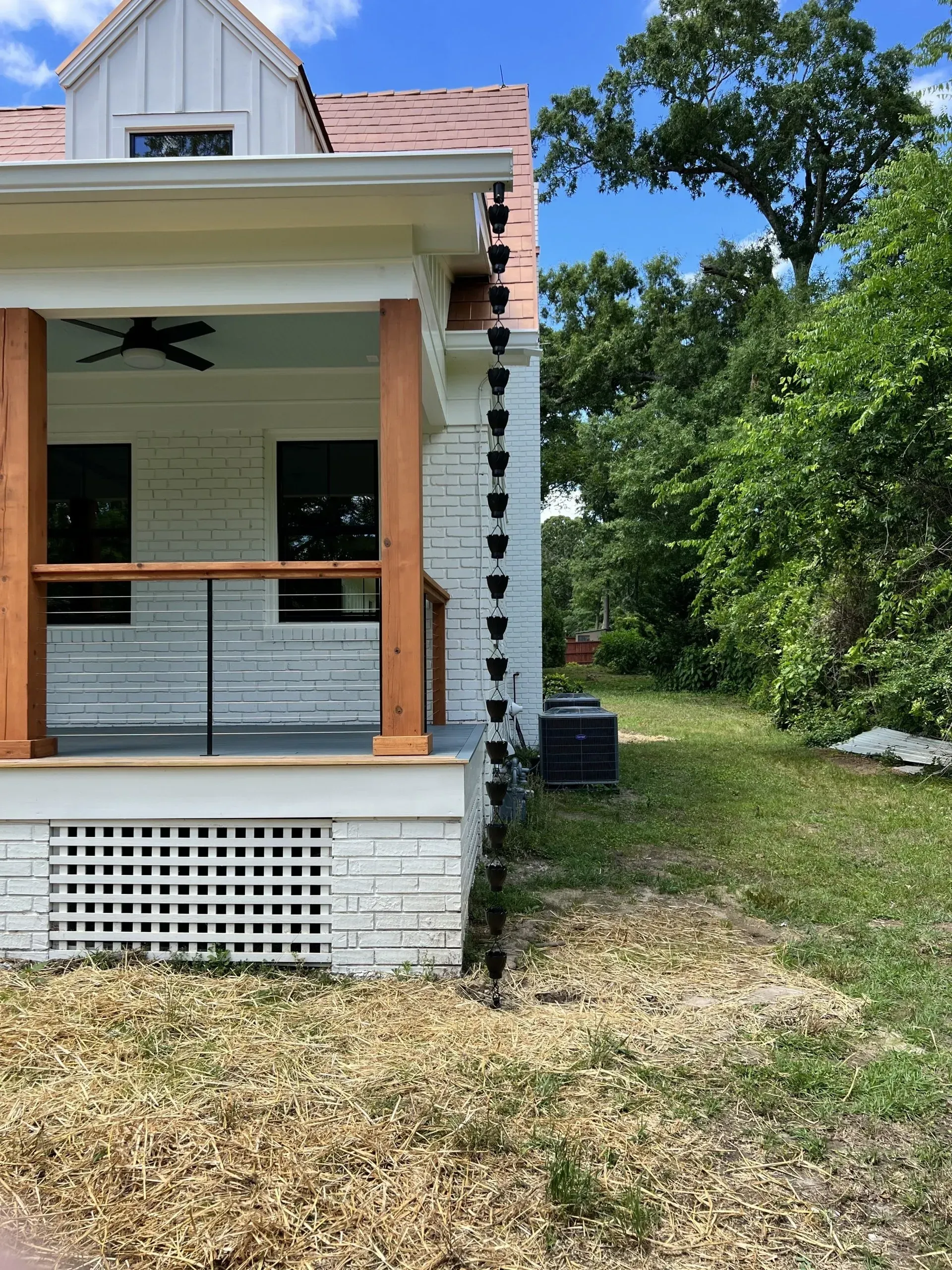 A house with a rain chain. White brick exterior, wooden porch with a black ceiling fan, and lush green trees.