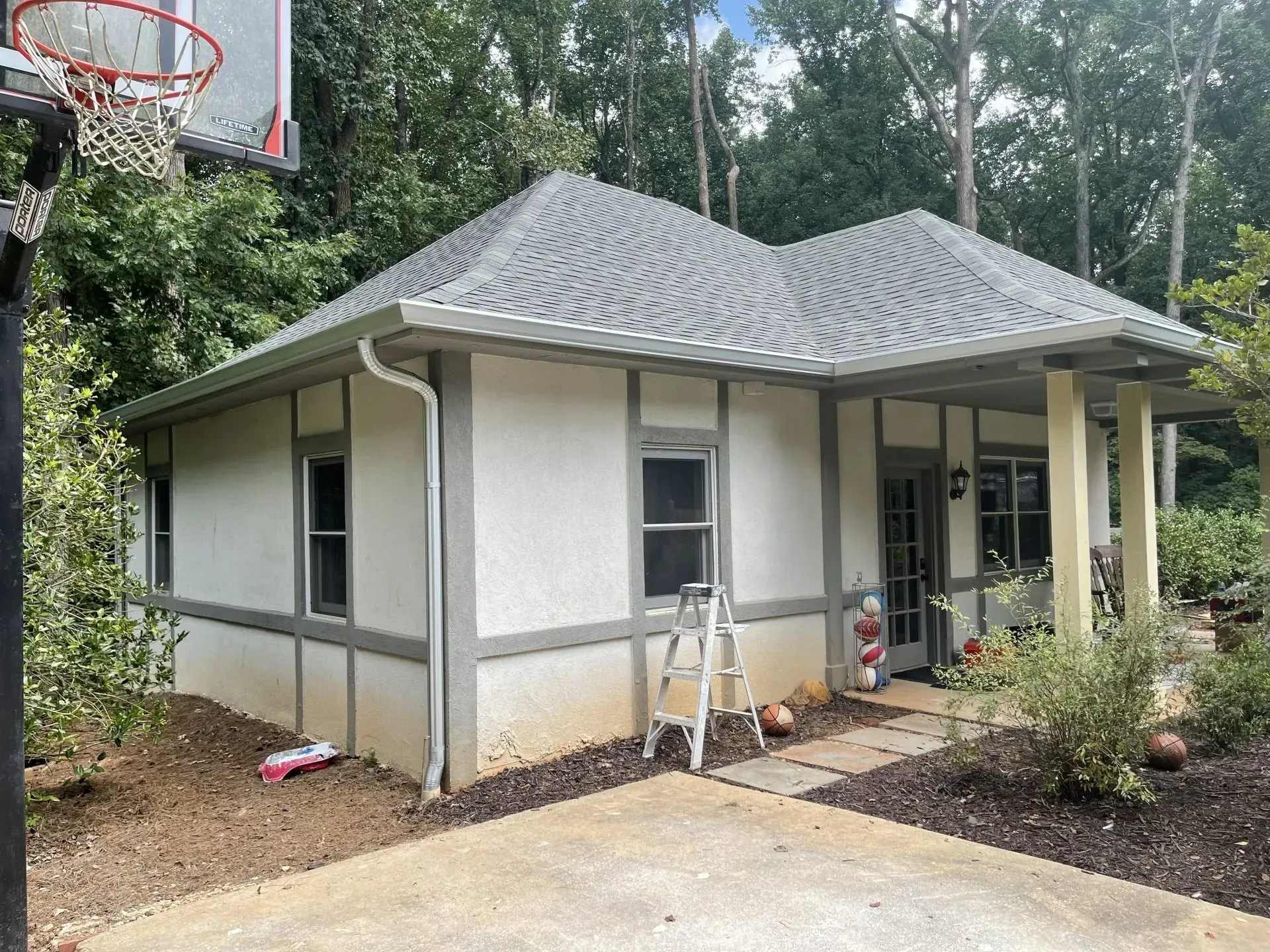 Small, white stucco house with gray trim and roof. A basketball hoop is visible.