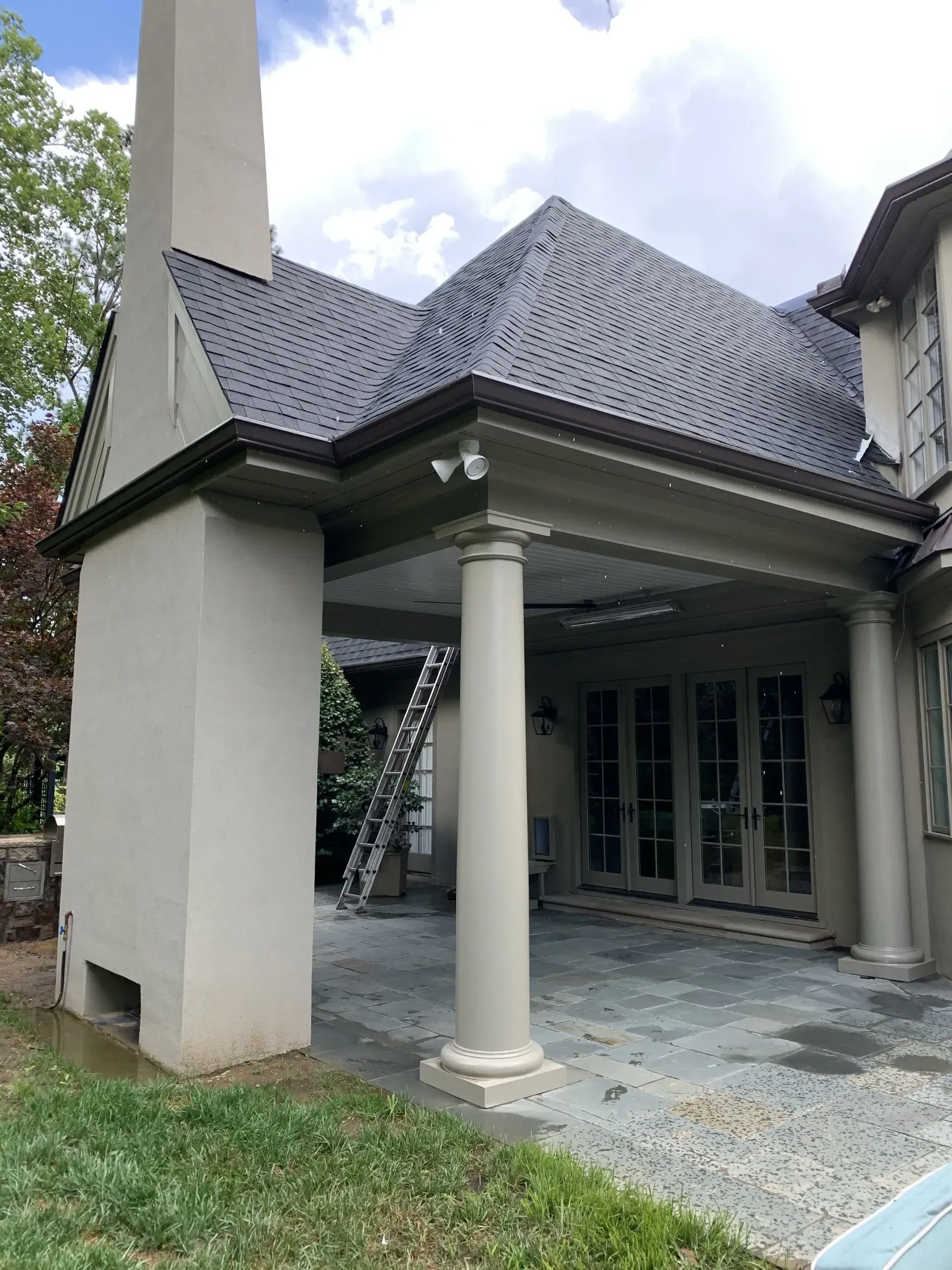 Covered patio with gray pillars, gray stone floor, and glass doors. Gray roof, chimney, and ladder visible.