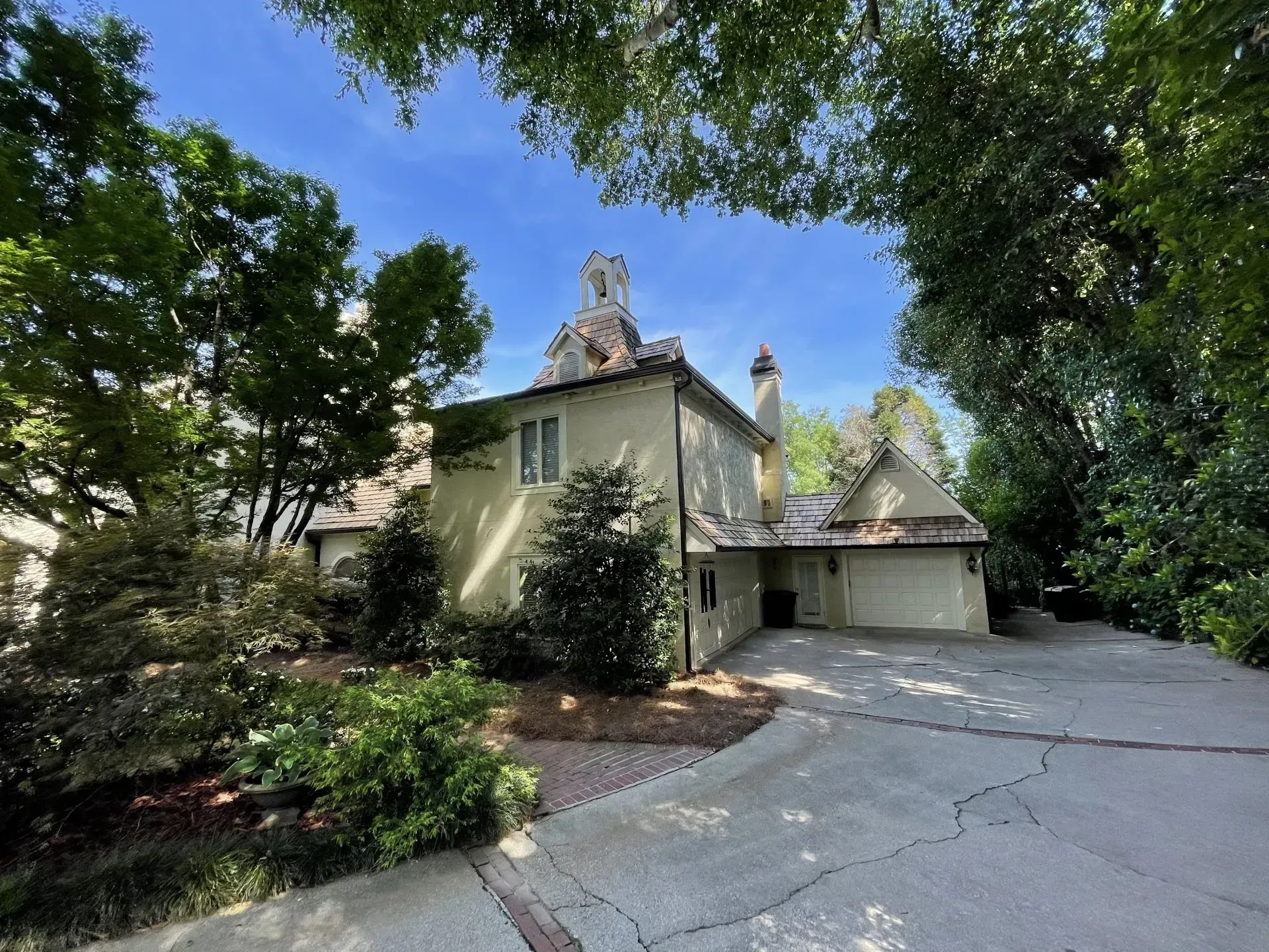 Beige house with a detached garage and driveway, surrounded by trees under a blue sky.