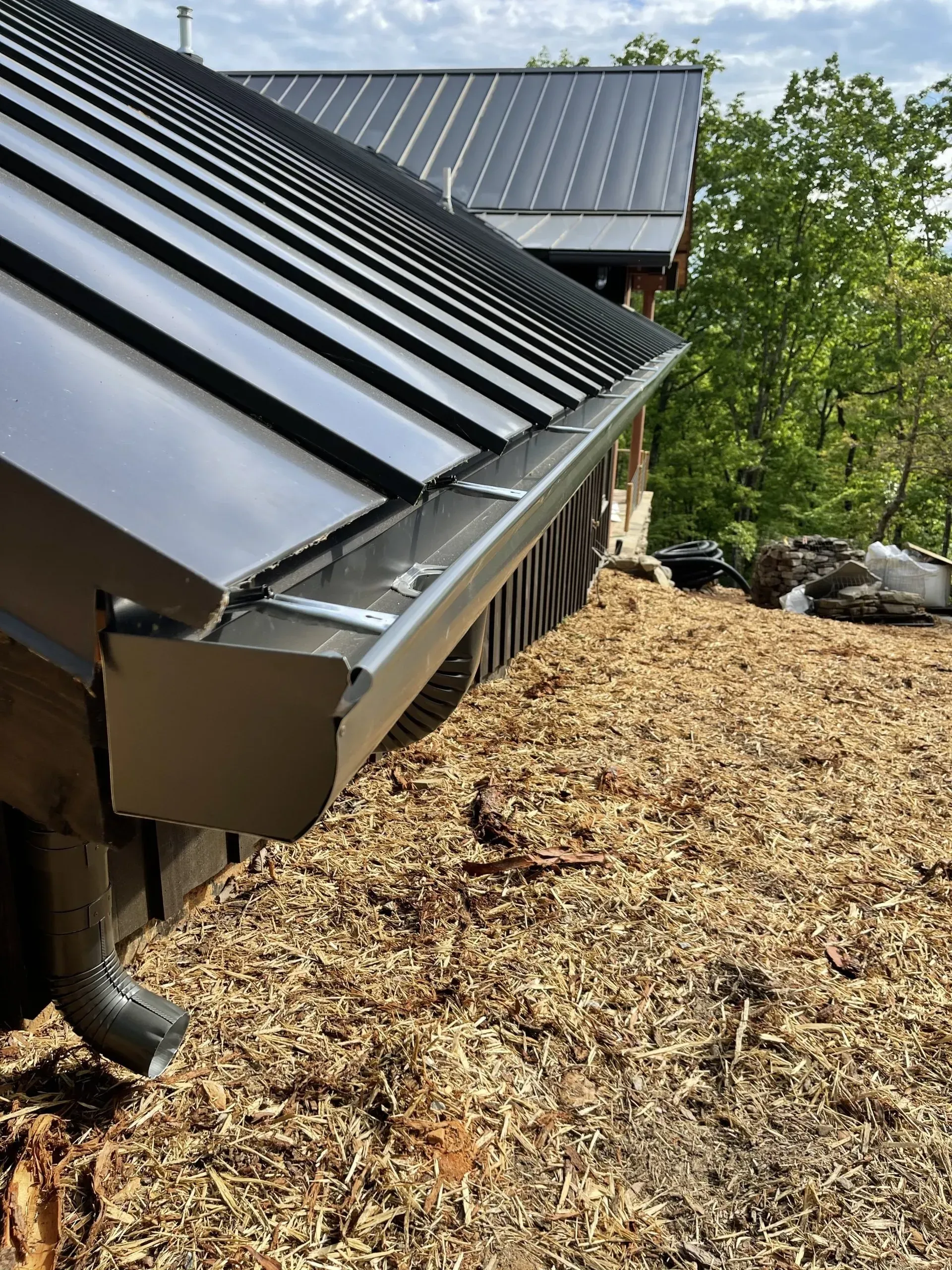 Black metal roof with matching gutters and a downspout against a wood chip-covered ground.