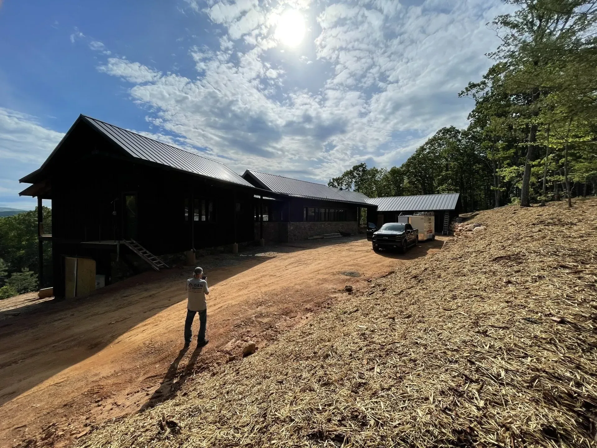 Dark house on a hillside with a dirt driveway and a person standing in front of it. Blue sky.