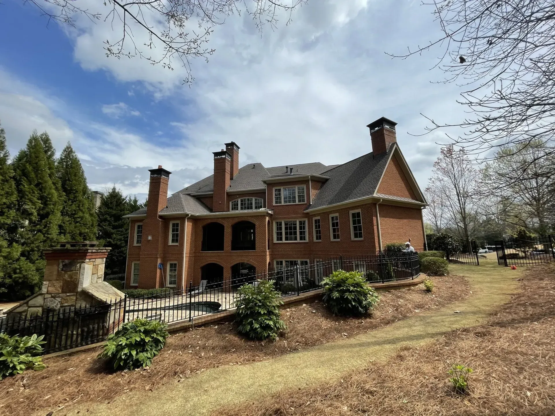 Large brick house with multiple chimneys and windows on a grassy hill, under a partly cloudy sky.