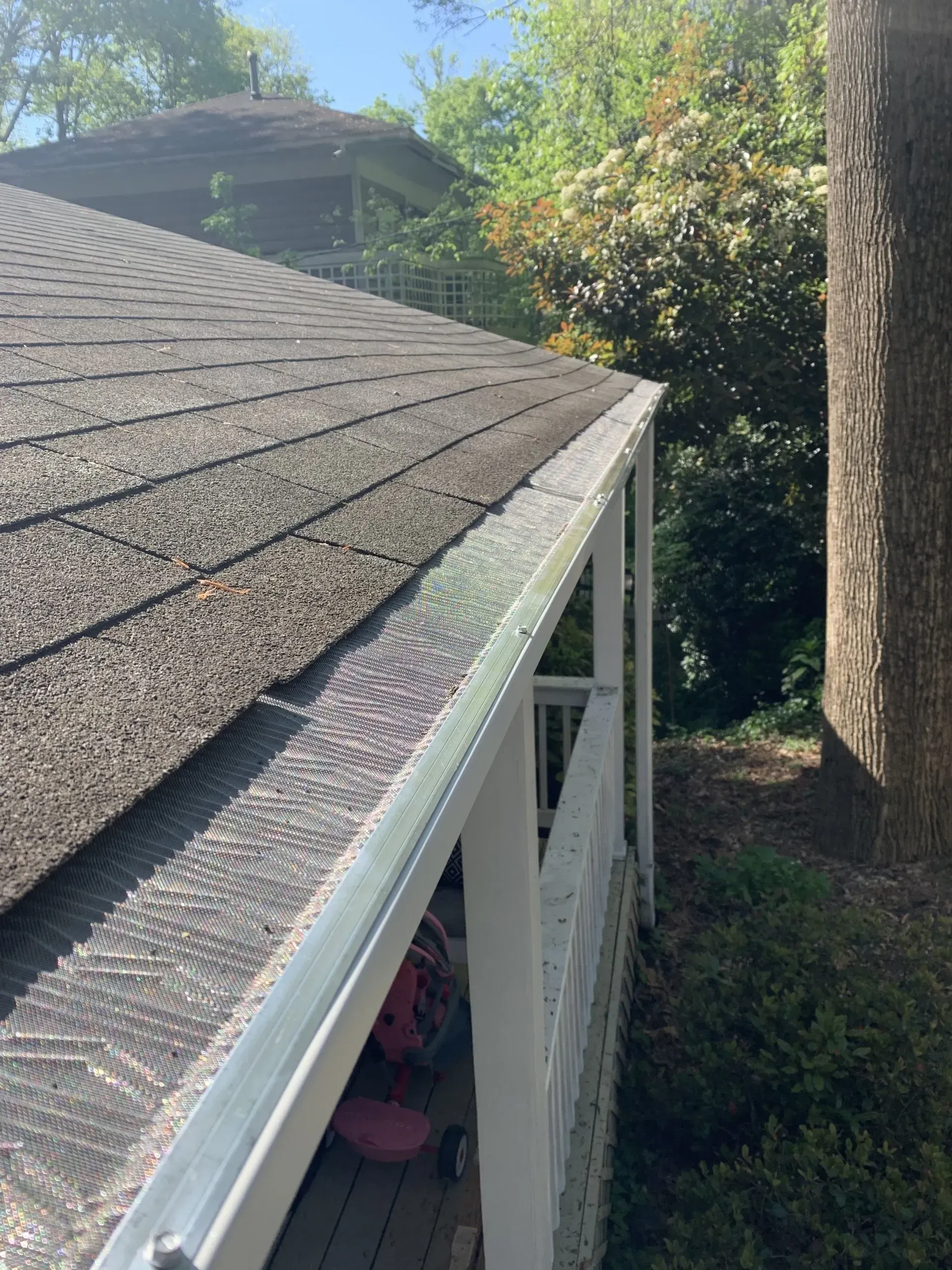 Gutter with leaf guard on a roof next to a white porch with trees in the background.