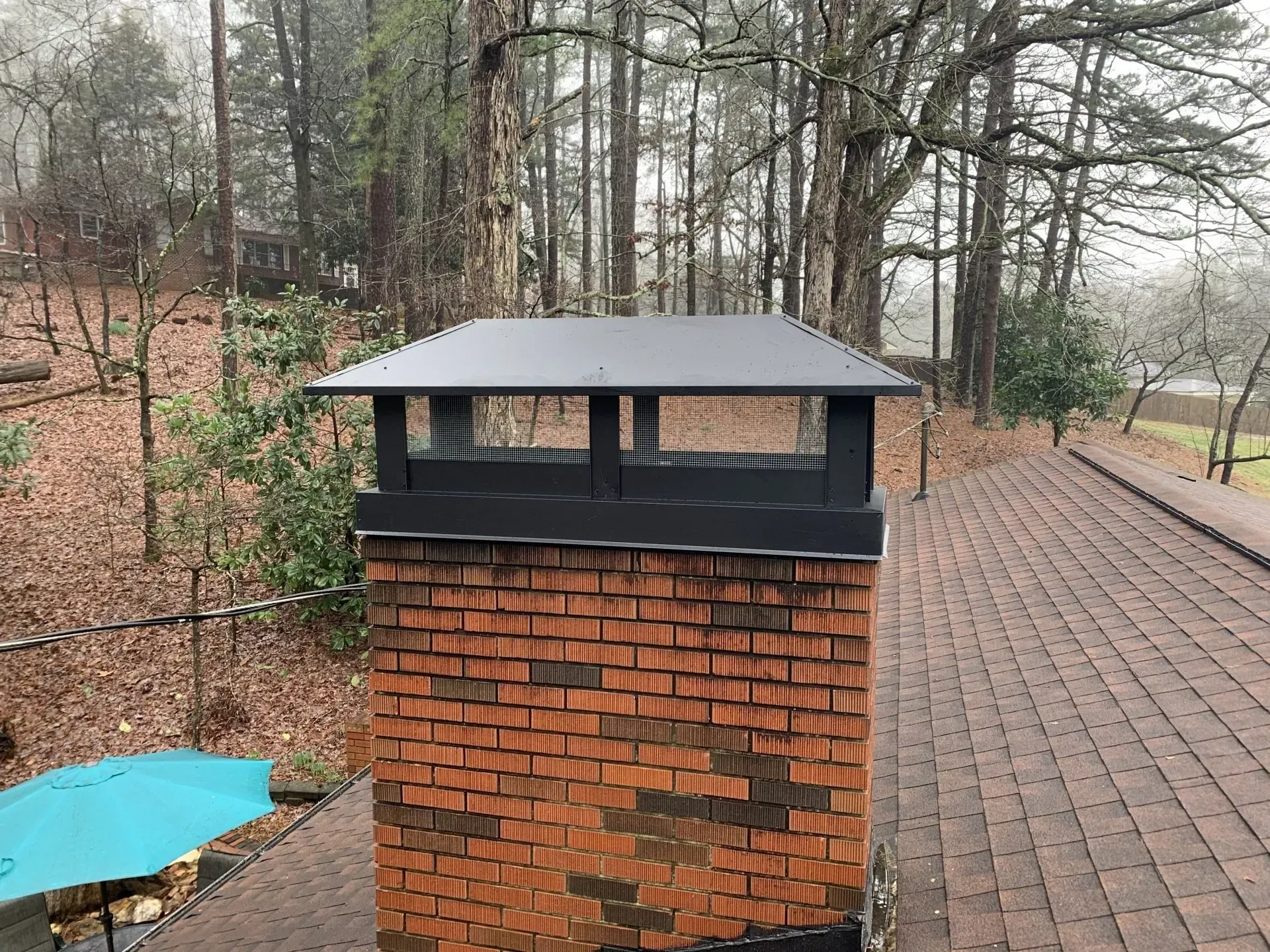 Brick chimney with a black metal chimney cap on a brown shingled roof, trees in the background.