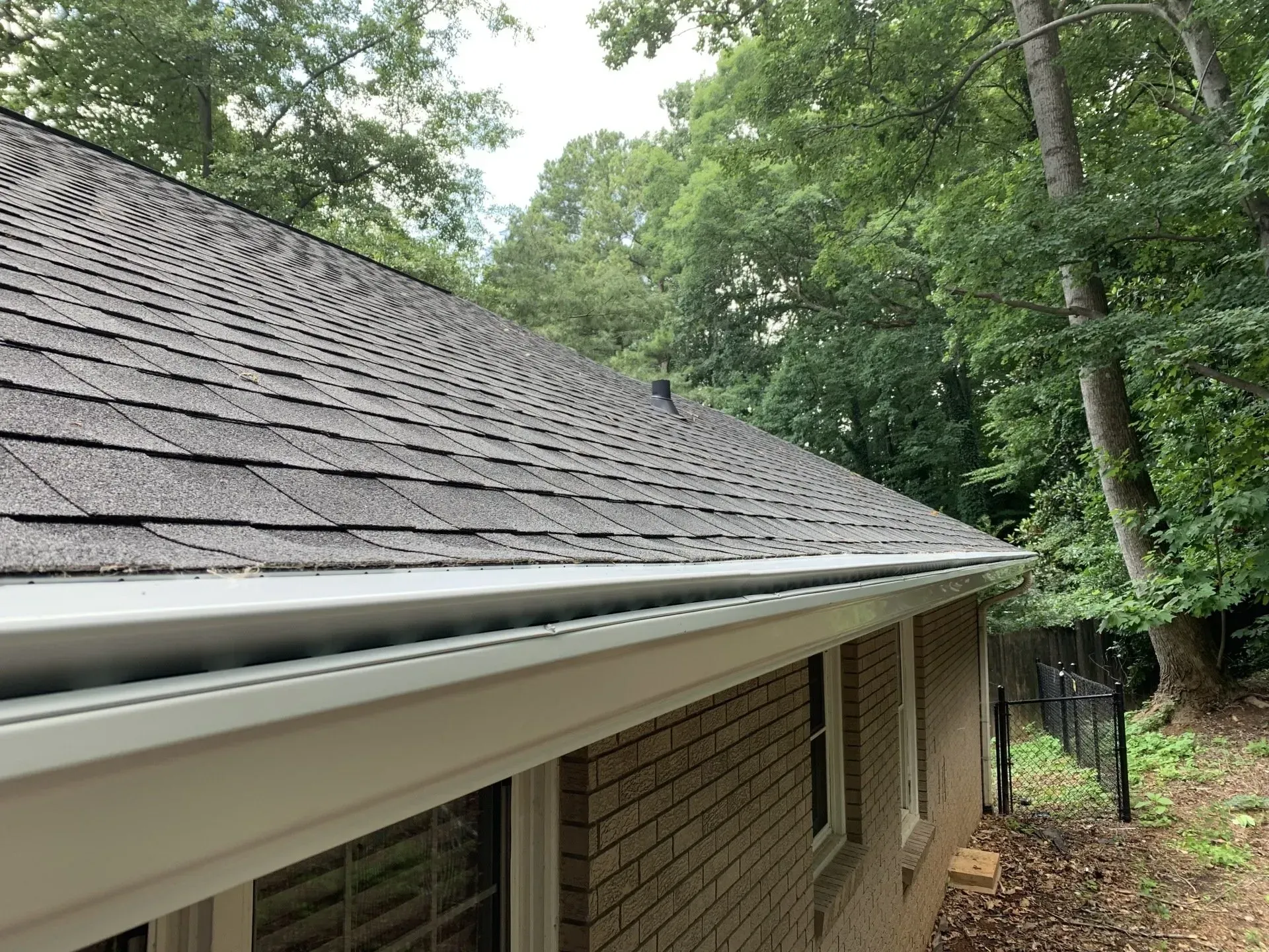 Gray shingle roof with white gutters on a brick home, surrounded by green trees.