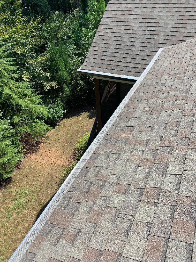 Overhead view of two shingled roofs, one light brown, the other dark brown. Lush green trees are nearby.