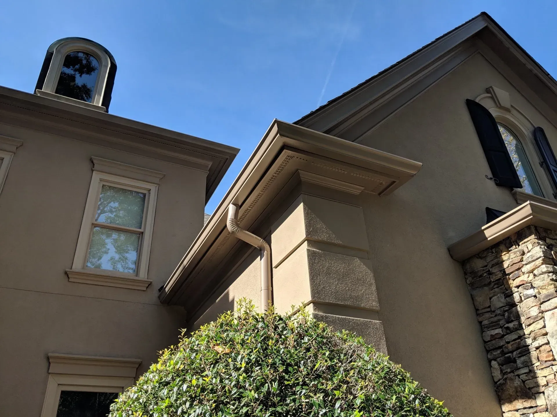 Beige building with brown roof and gutters against a blue sky, some windows and a stone accent.