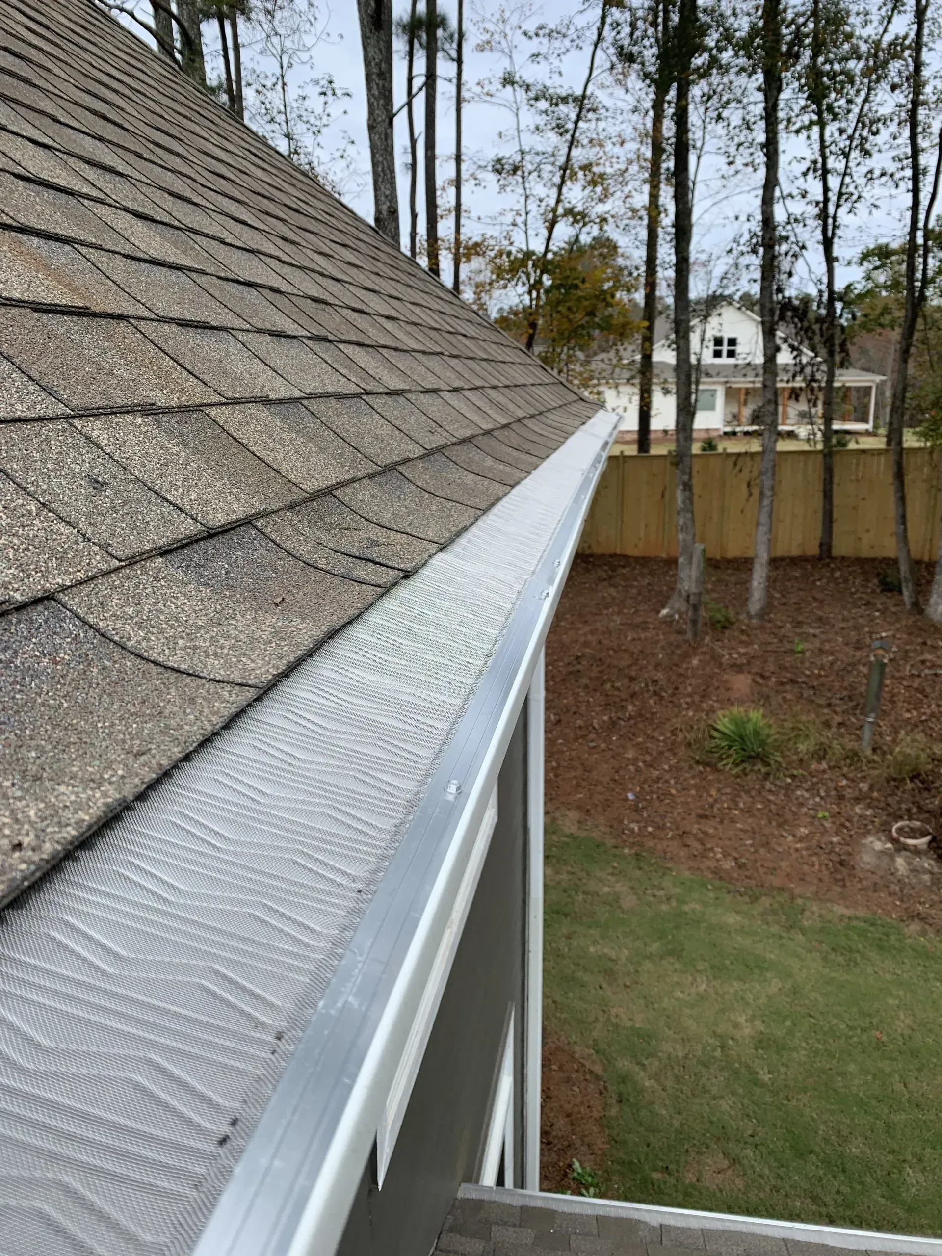 Close-up of a roof with shingle tiles and gutter with a mesh guard, surrounded by trees and a yard.