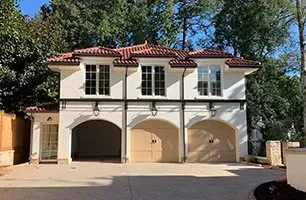 Three-car garage with arched entryways, windows above, and a red tile roof. Cream-colored exterior with a surrounding green trees.