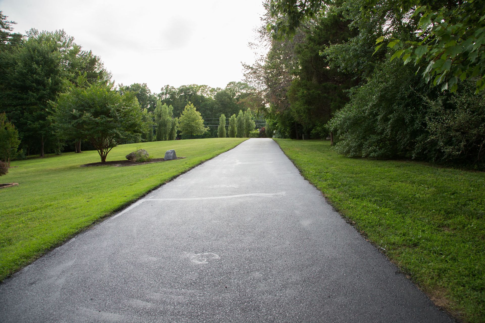 Asphalt driveway flanked by green lawns and trees, leading to a lighter horizon.