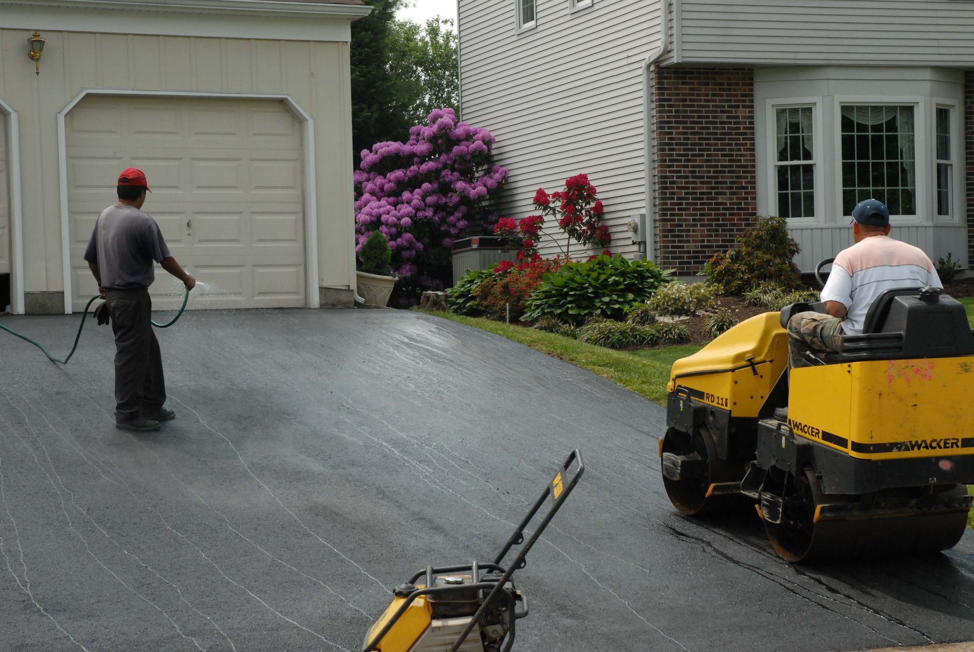 Two workers paving a driveway: one spraying, the other operating a yellow compactor roller. Houses in the background.
