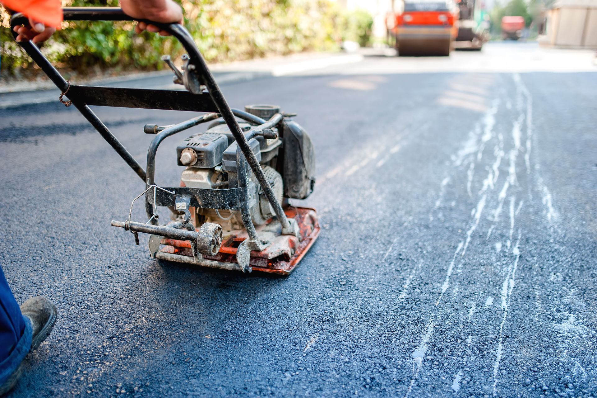 Person using a plate compactor to flatten fresh asphalt on a road.