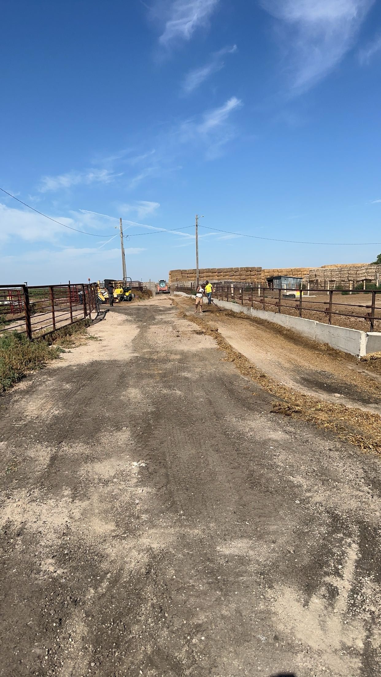 Dirt road leads to construction site under blue sky. A tractor and stacked lumber are visible.