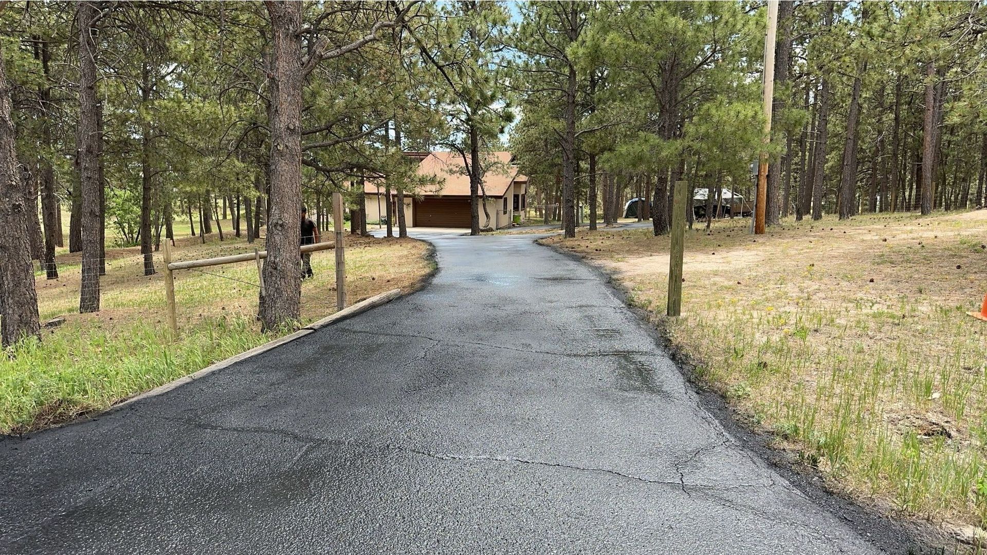 Asphalt driveway leading to a brown house through a wooded area. Trees line both sides.