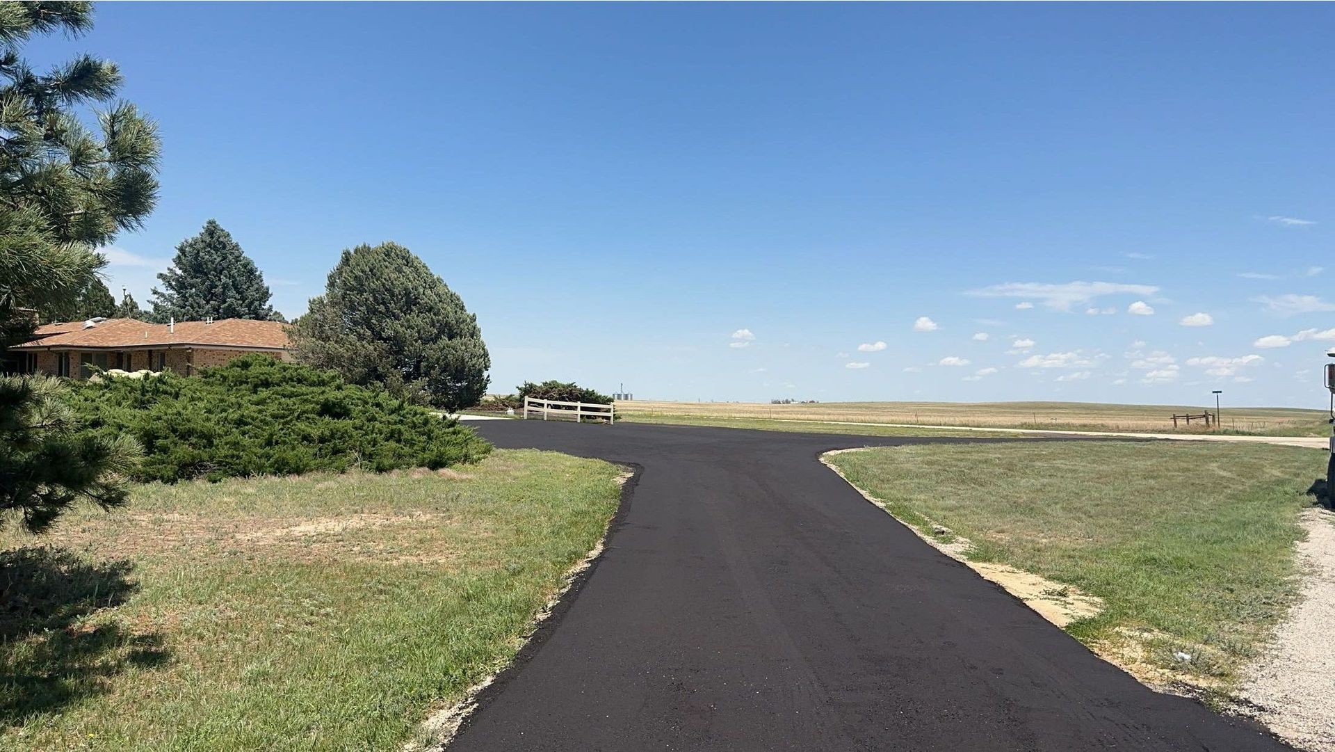 A paved road forks towards a house and an open field under a blue sky.