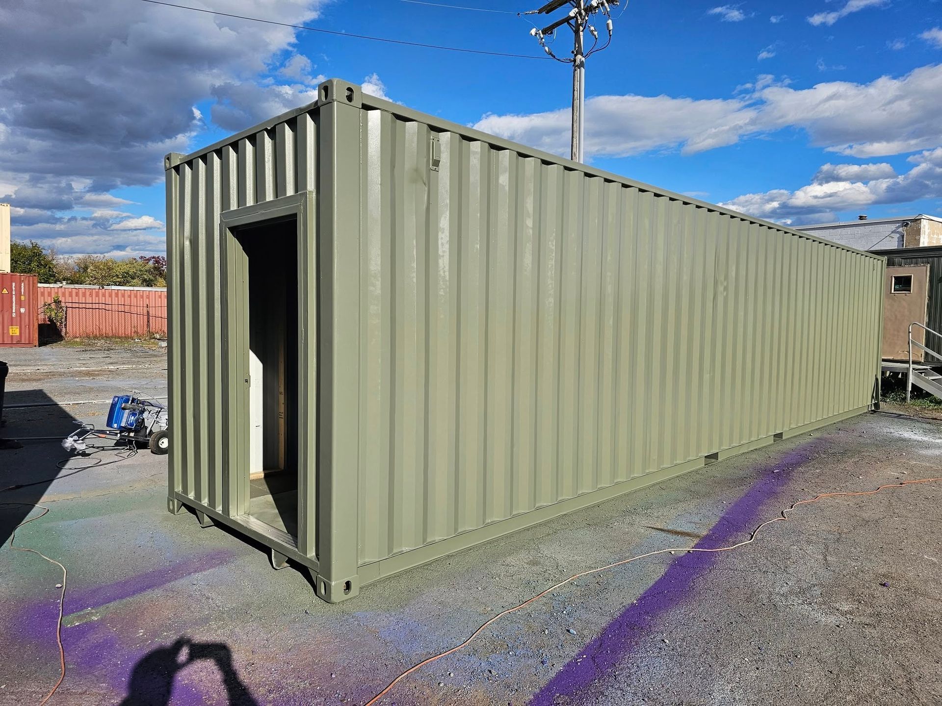 A long, olive green industrial shipping container with an open doorway, sitting on a gravel lot under a sunny sky.
