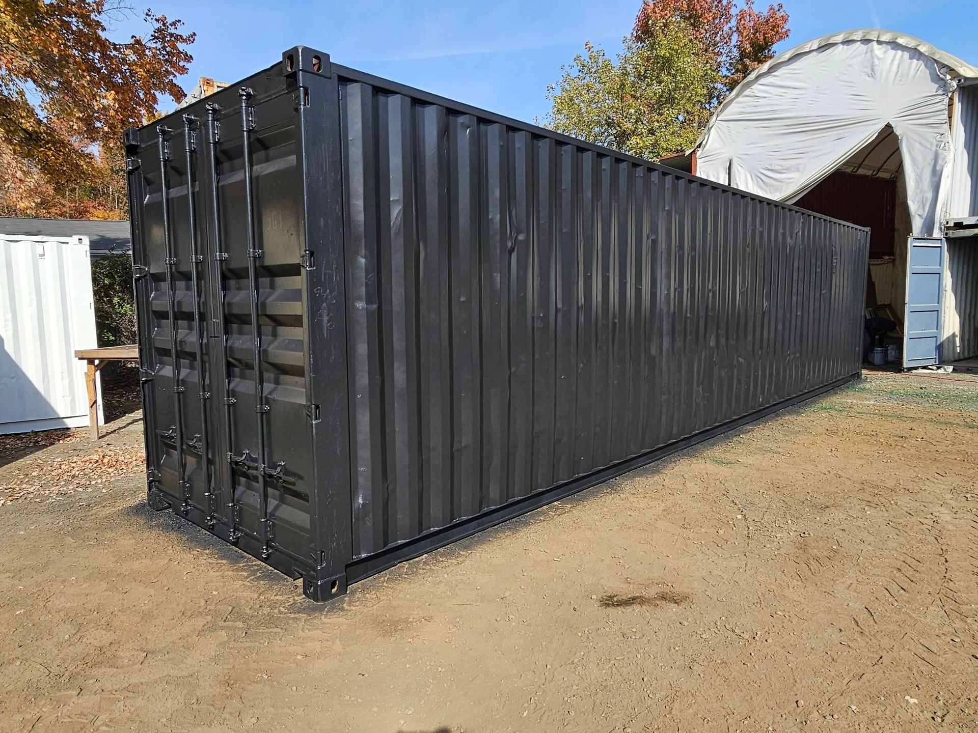 A black 40-foot shipping container stands on a gravel lot under a clear blue sky.