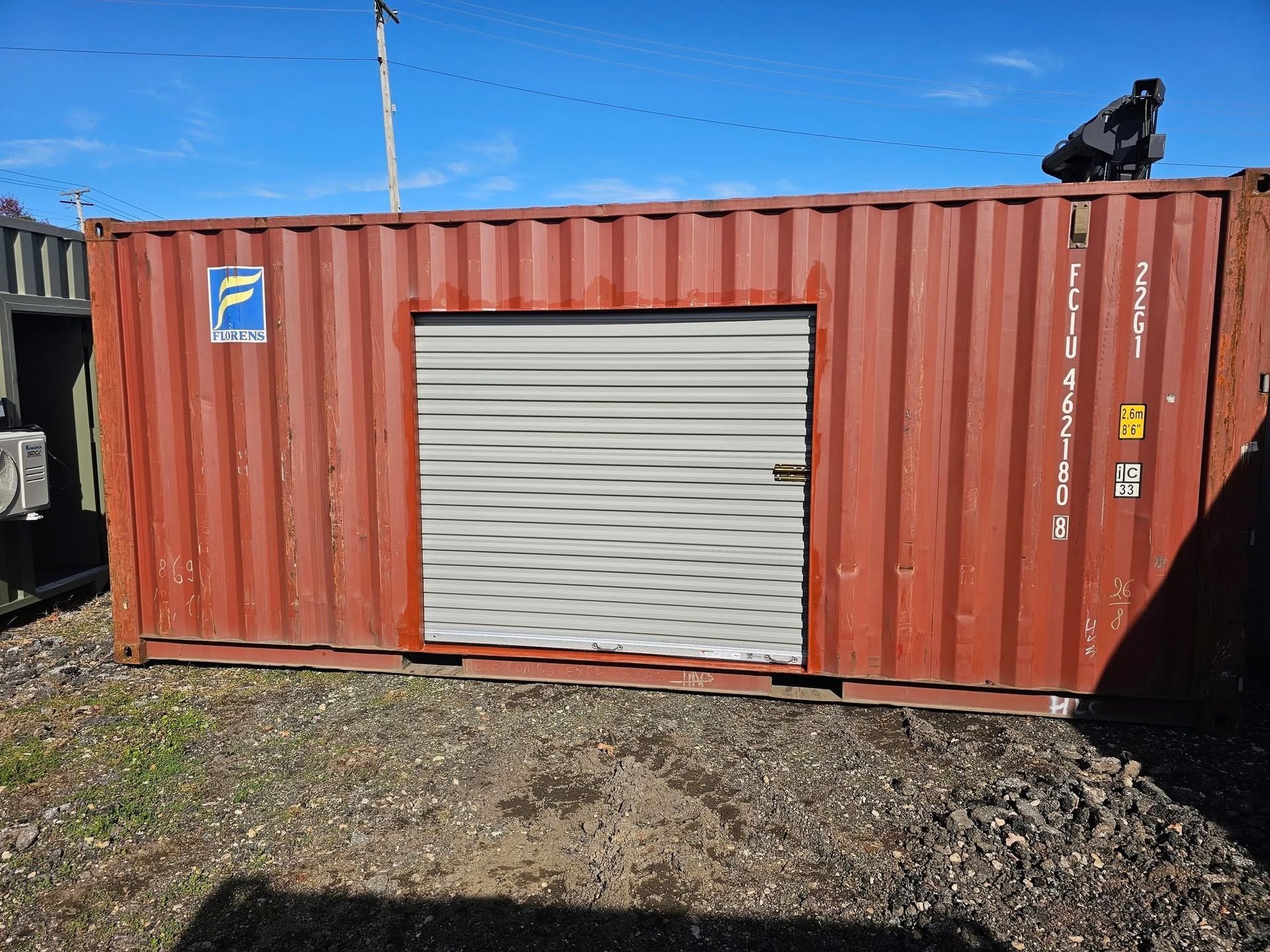 A rusted red shipping container with a centered, beige roll-up garage door sitting on gravel under a blue sky.