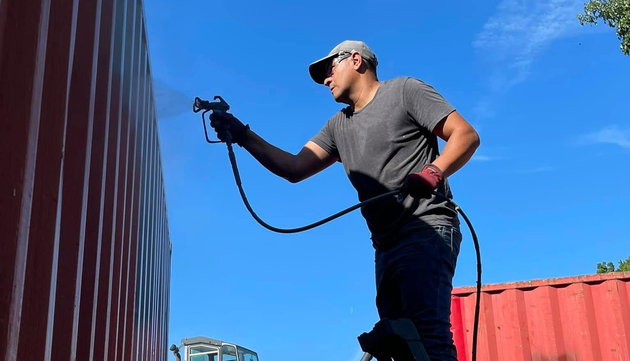A person wearing protective gear uses a paint sprayer on a red shipping container against a clear blue sky.