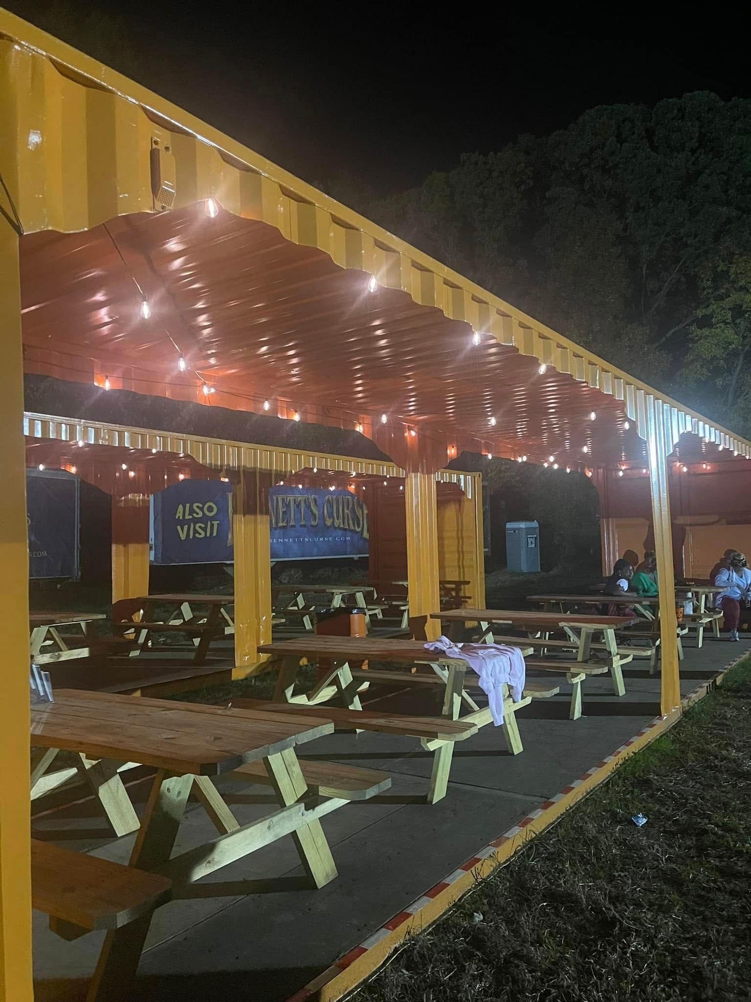 Picnic tables sit under a yellow outdoor shelter illuminated by string lights at night.