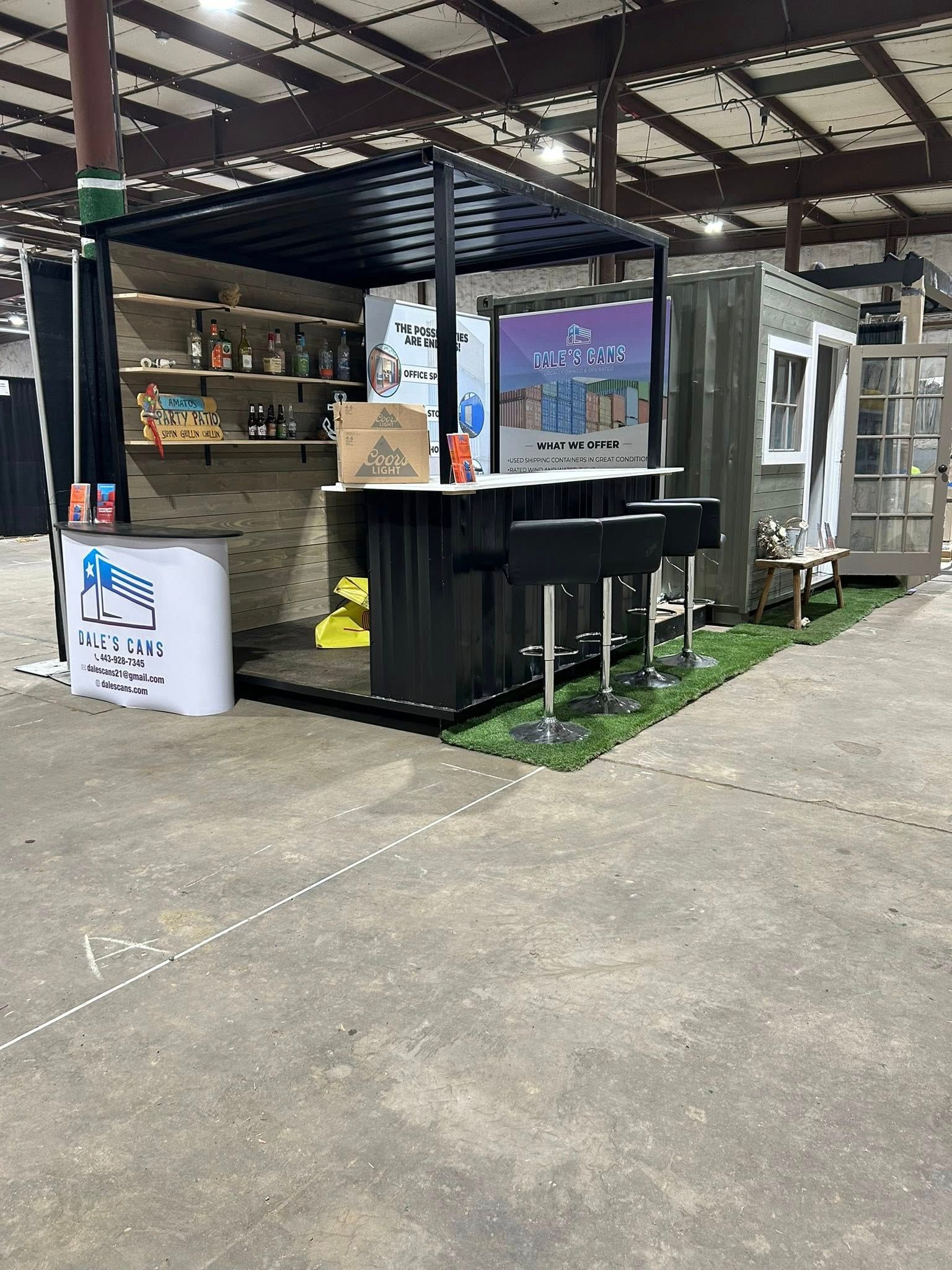 A portable bar booth with a black metal frame, shelves, stools, and signage, set up on a patch of artificial grass indoors.