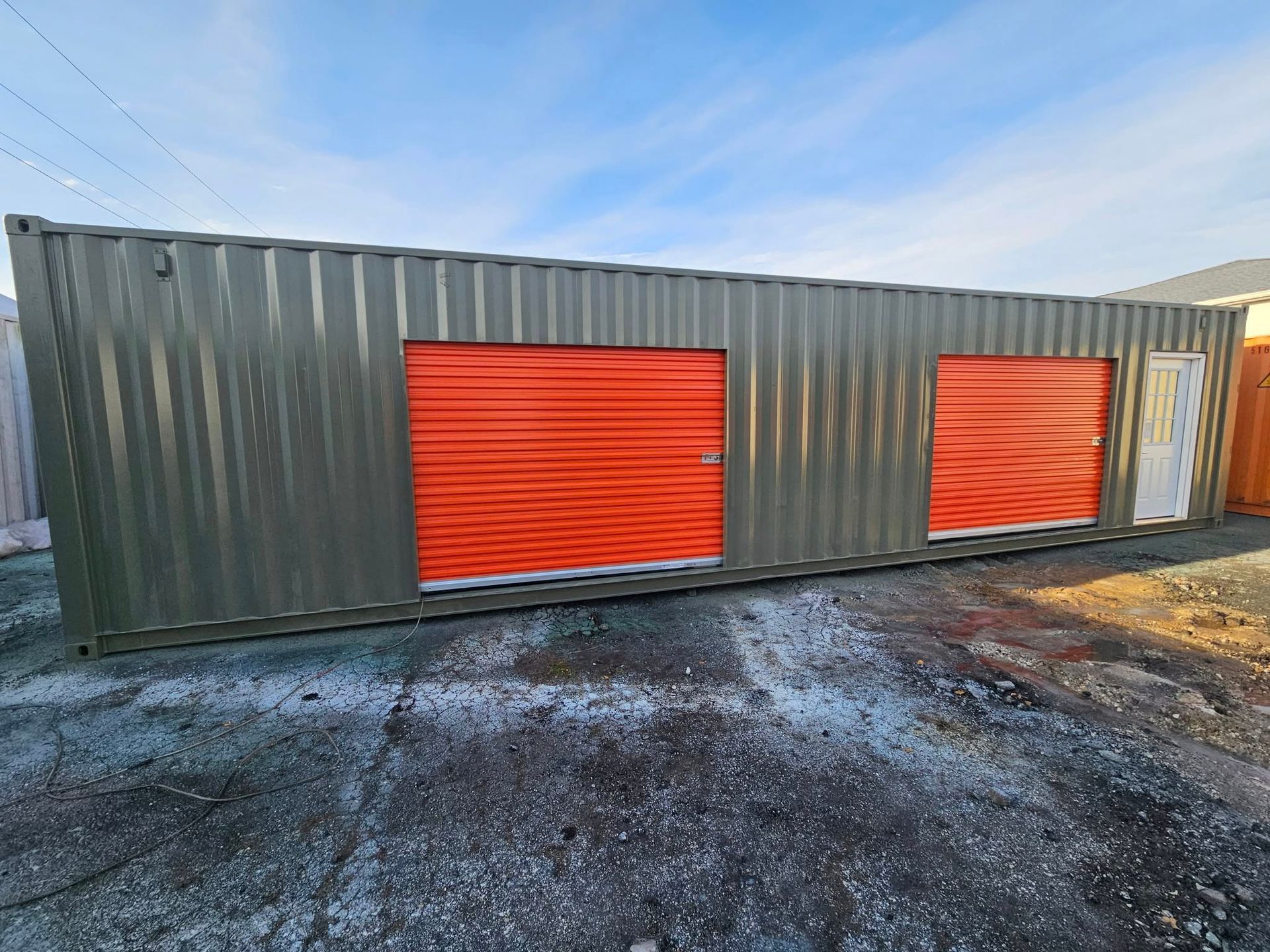 An olive-green shipping container modified into storage units with two orange roll-up doors and a white pedestrian door.