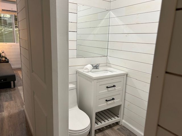 A small bathroom featuring white shiplap walls, a white vanity with two drawers and an open shelf, and a white toilet.