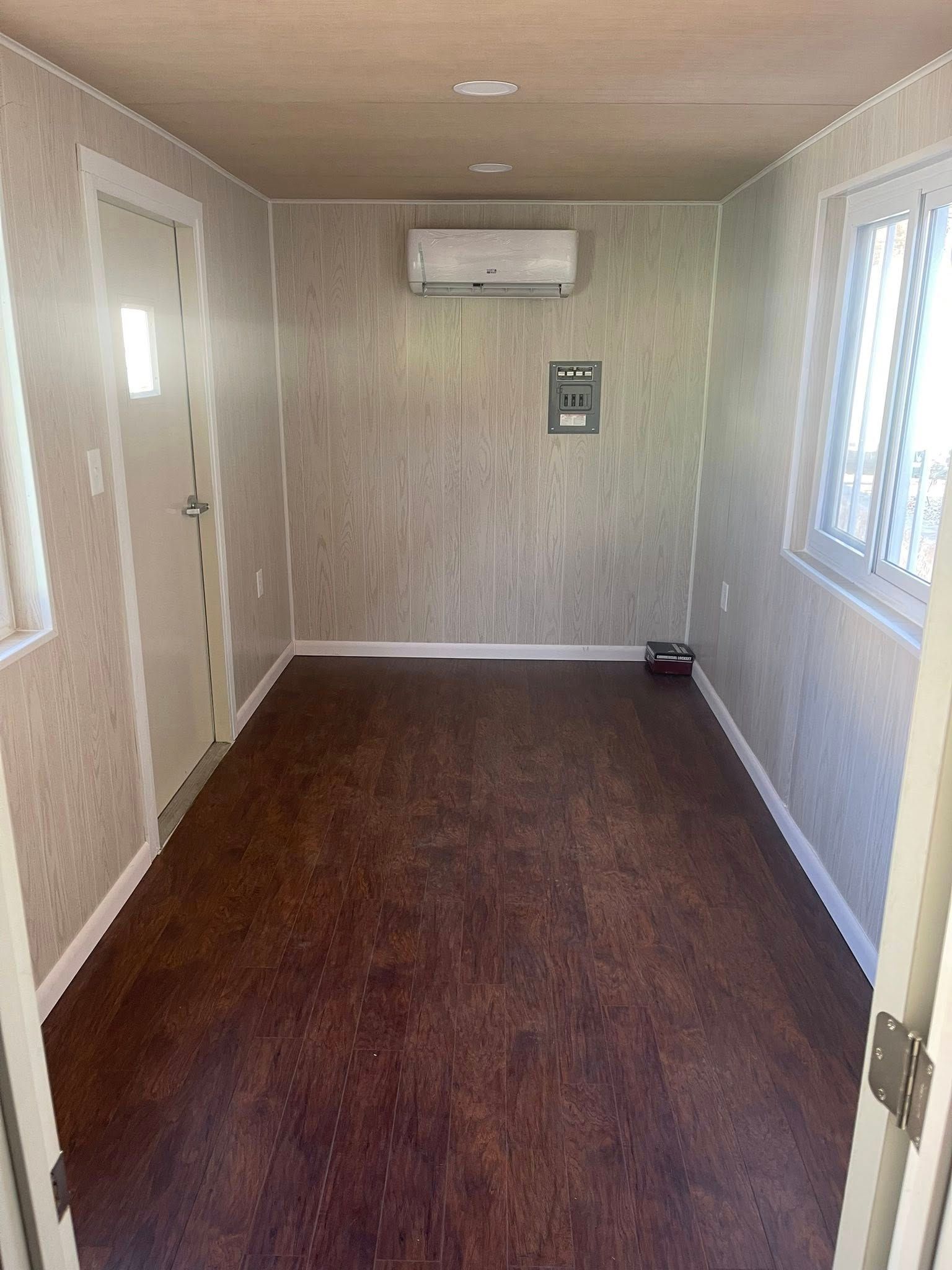 A small, empty room with wood-patterned walls, dark flooring, a white door, a window, and a wall-mounted AC unit.