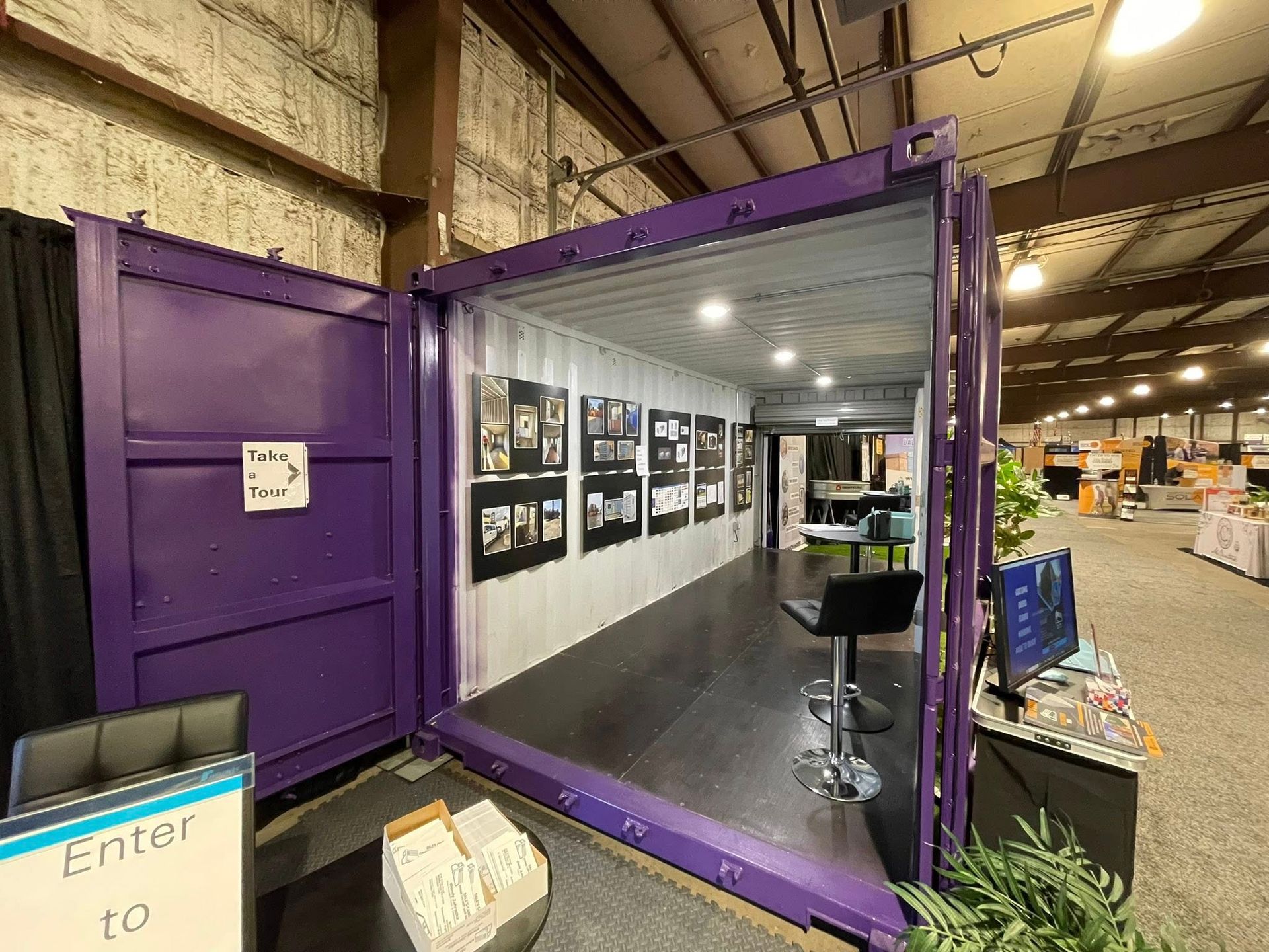 A purple shipping container converted into an indoor display booth with framed photos on the wall and a stool inside.