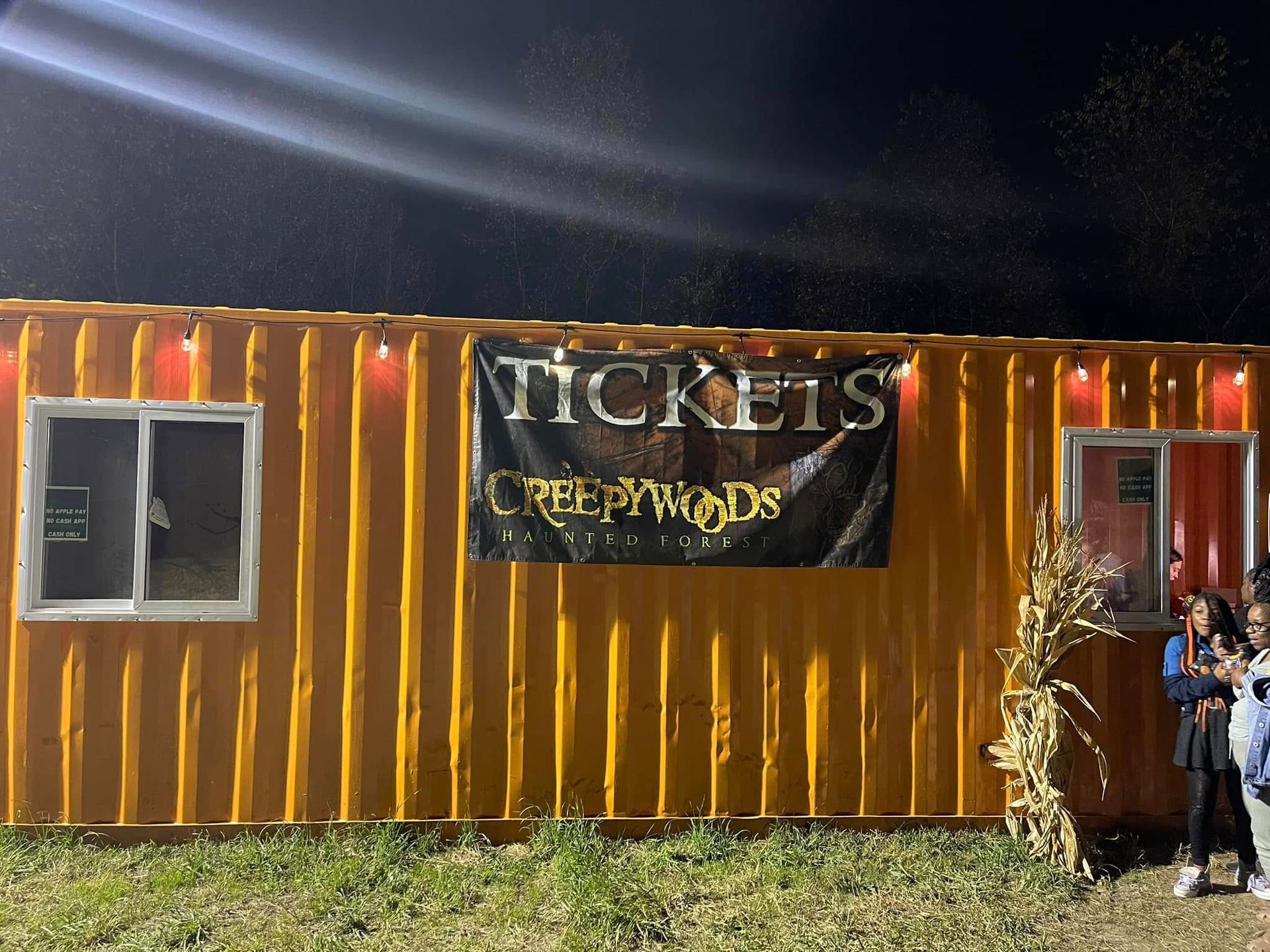 A yellow container serving as a ticket booth for Creepywoods, decorated with string lights and a bundle of dried corn.