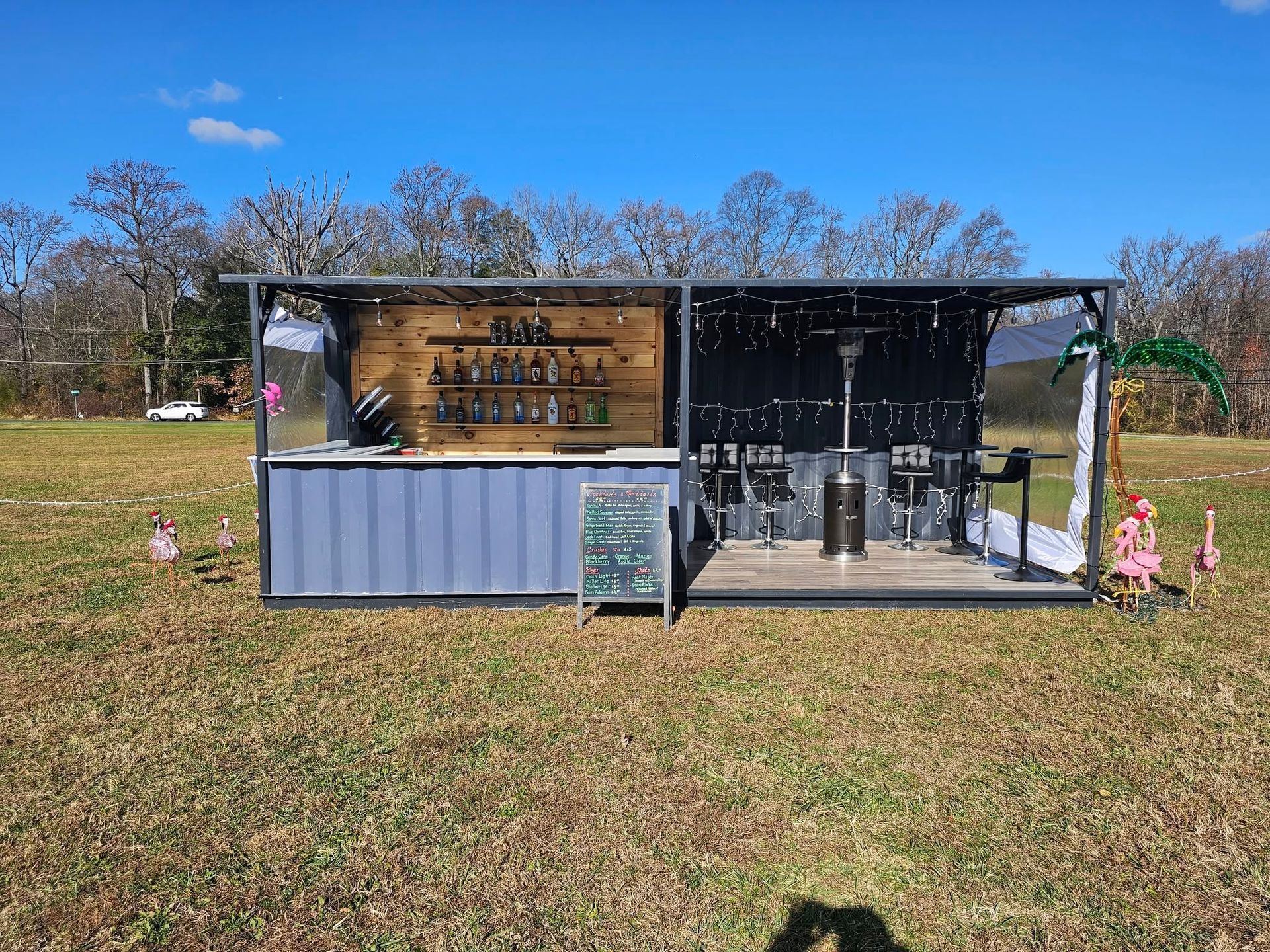 A modern outdoor bar built into a gray shipping container, set in a sunny field with decorative flamingo figures.