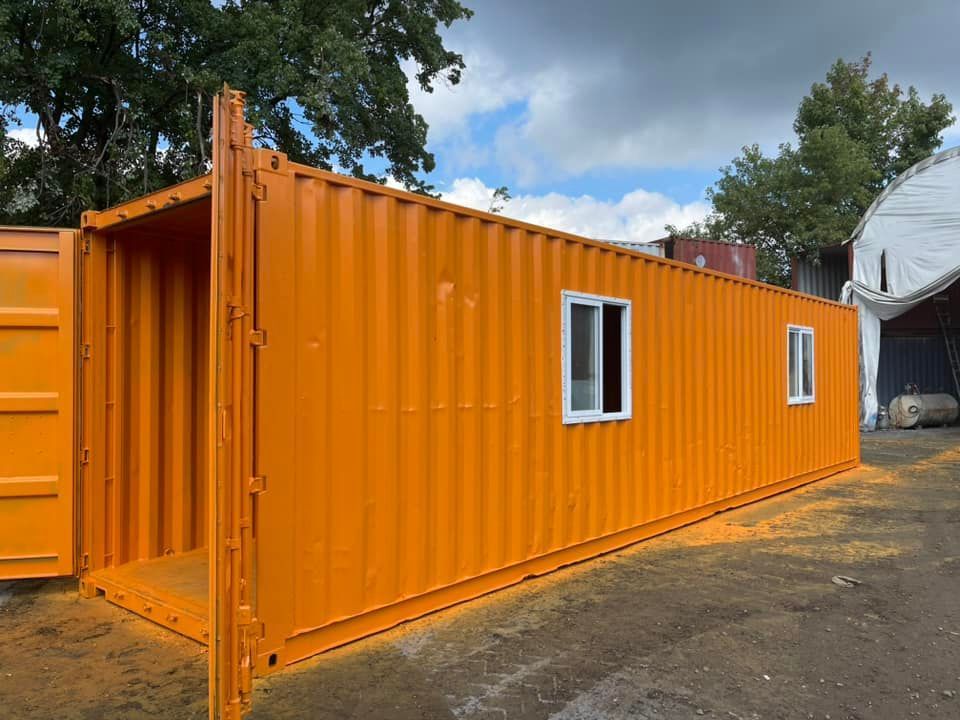 A bright orange repurposed shipping container with two windows, standing outdoors on a gravel lot.