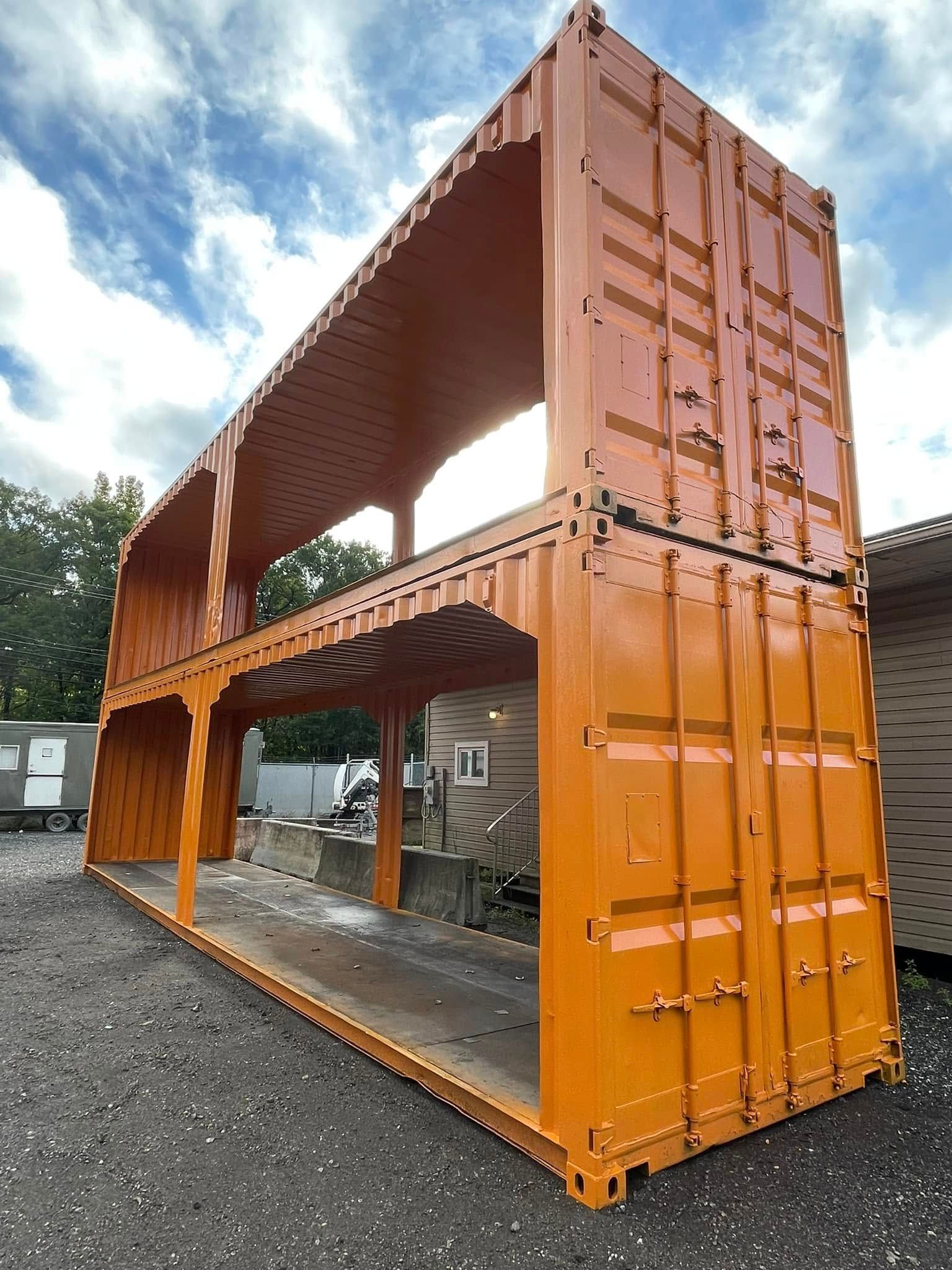 Two stacked bright orange shipping containers with their side walls removed, standing on a gravel surface.