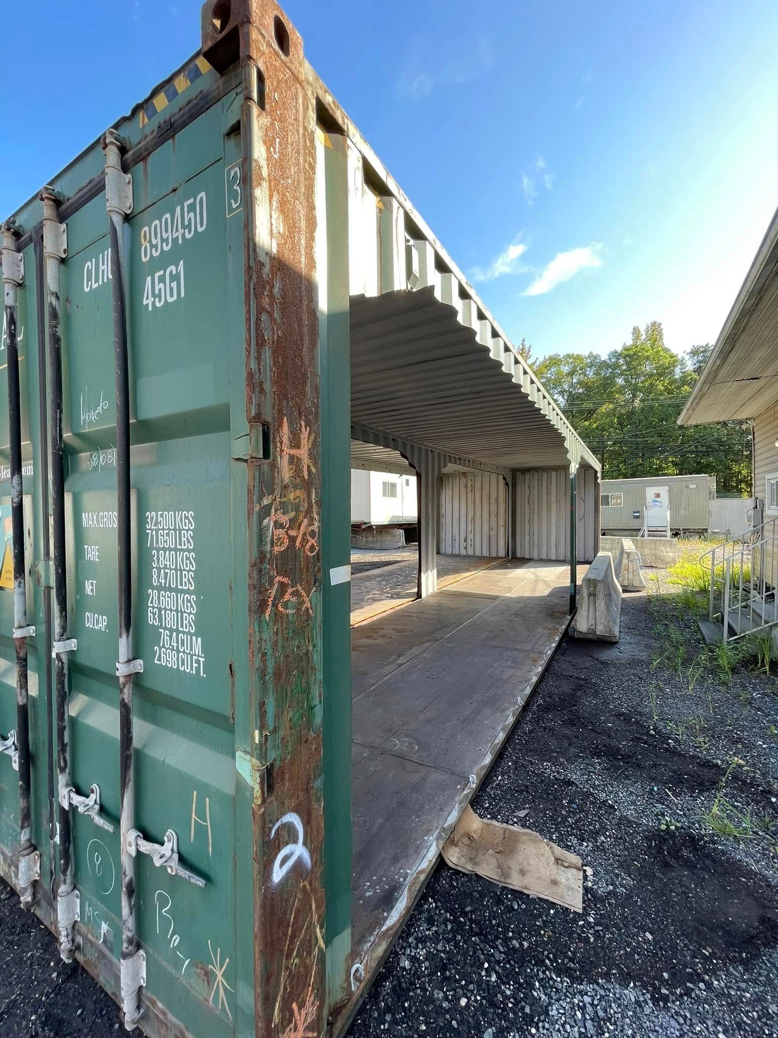 A green, open-ended shipping container structure stands on a gravel lot under a bright blue sky.