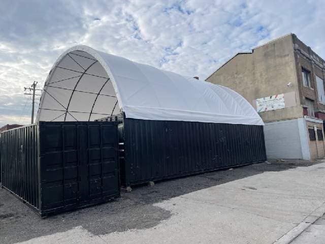 A white arched fabric shelter mounted on top of two black shipping containers in an outdoor lot next to a brick building.