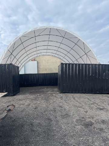 A large, white, arched fabric roof structure resting on two parallel rows of black shipping containers outdoors.