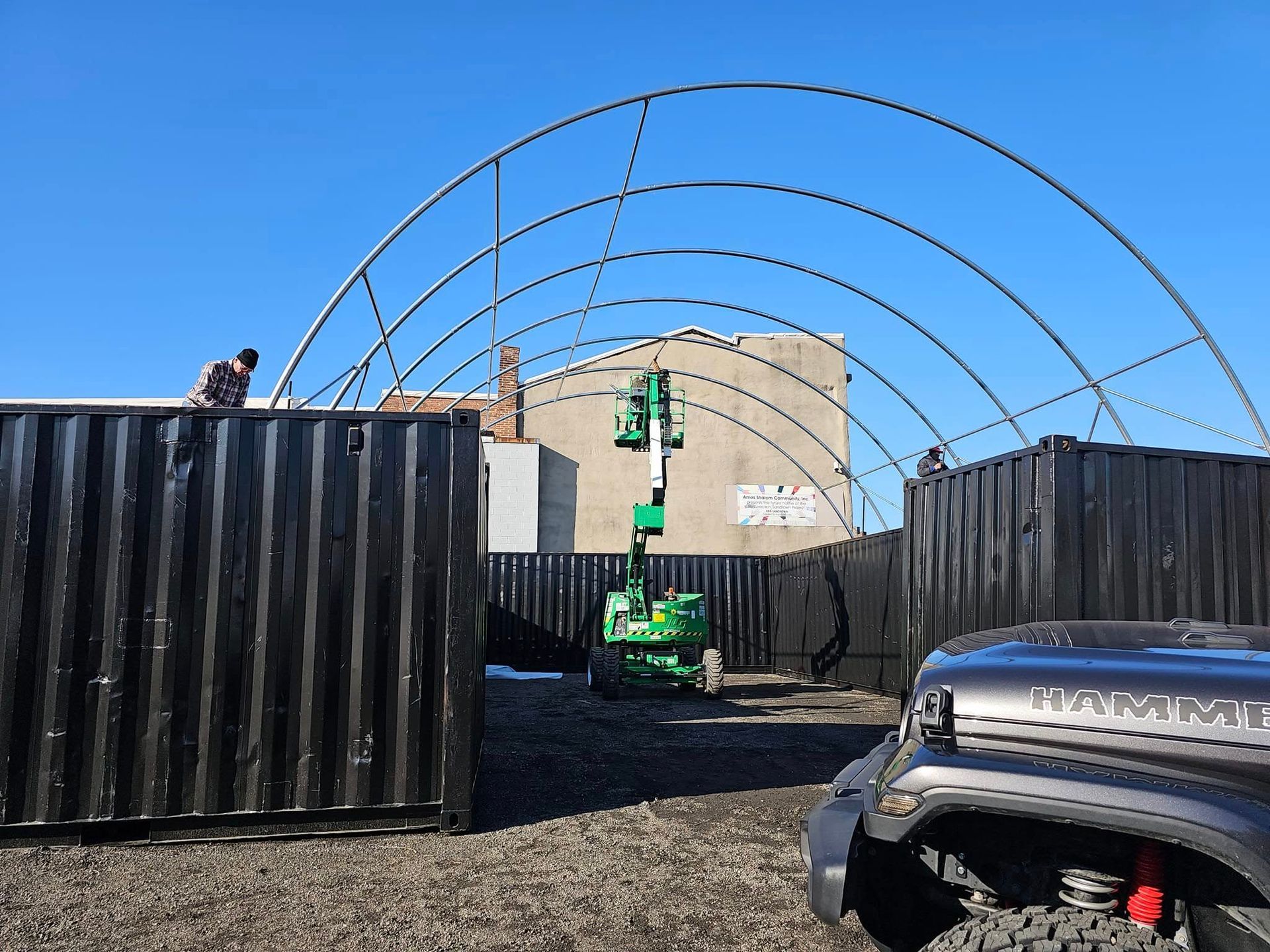 A construction site with a curved metal frame arching between two black shipping containers, with a green lift nearby.