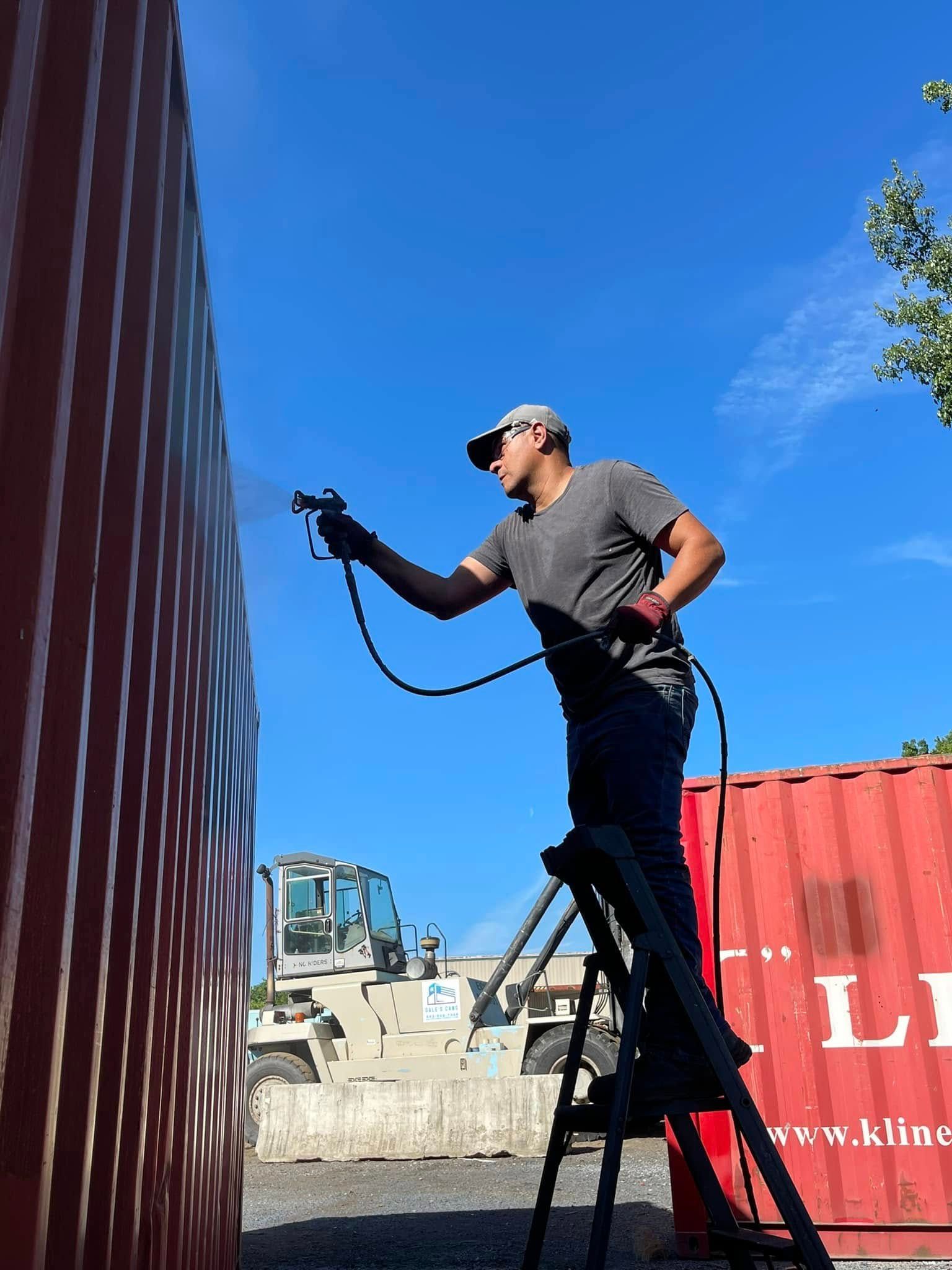 A person on a stepladder uses a paint sprayer on a red shipping container outdoors under a bright blue sky.