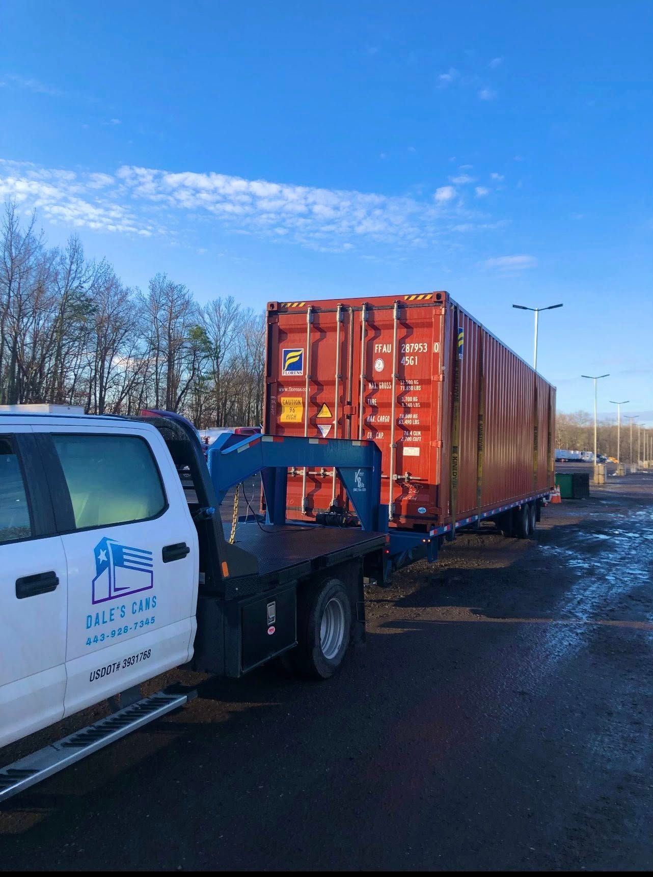 A white truck hitched to a flatbed trailer hauling a large, red shipping container on a muddy lot under a blue sky.