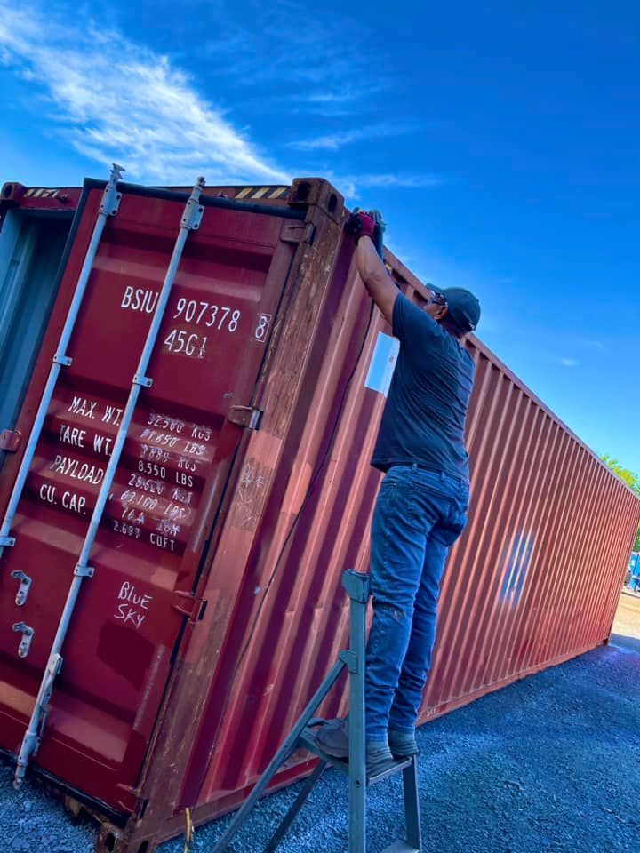 A person on a ladder reaches to attach a label to the side of a tall, red shipping container under a blue sky.