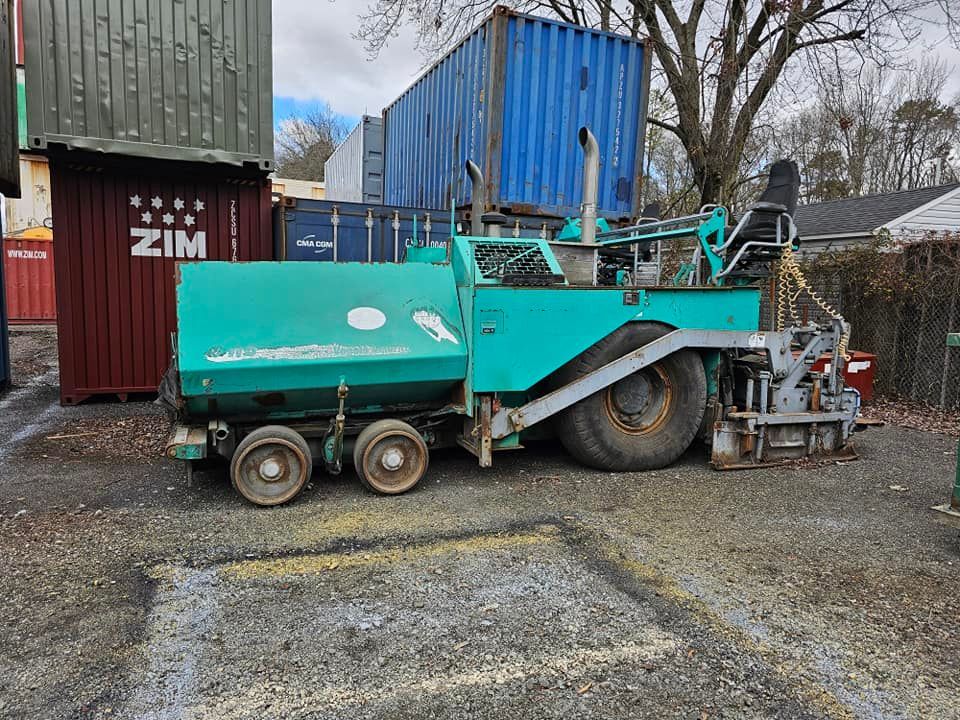 A teal asphalt paver parked on a gravel lot near shipping containers and trees.