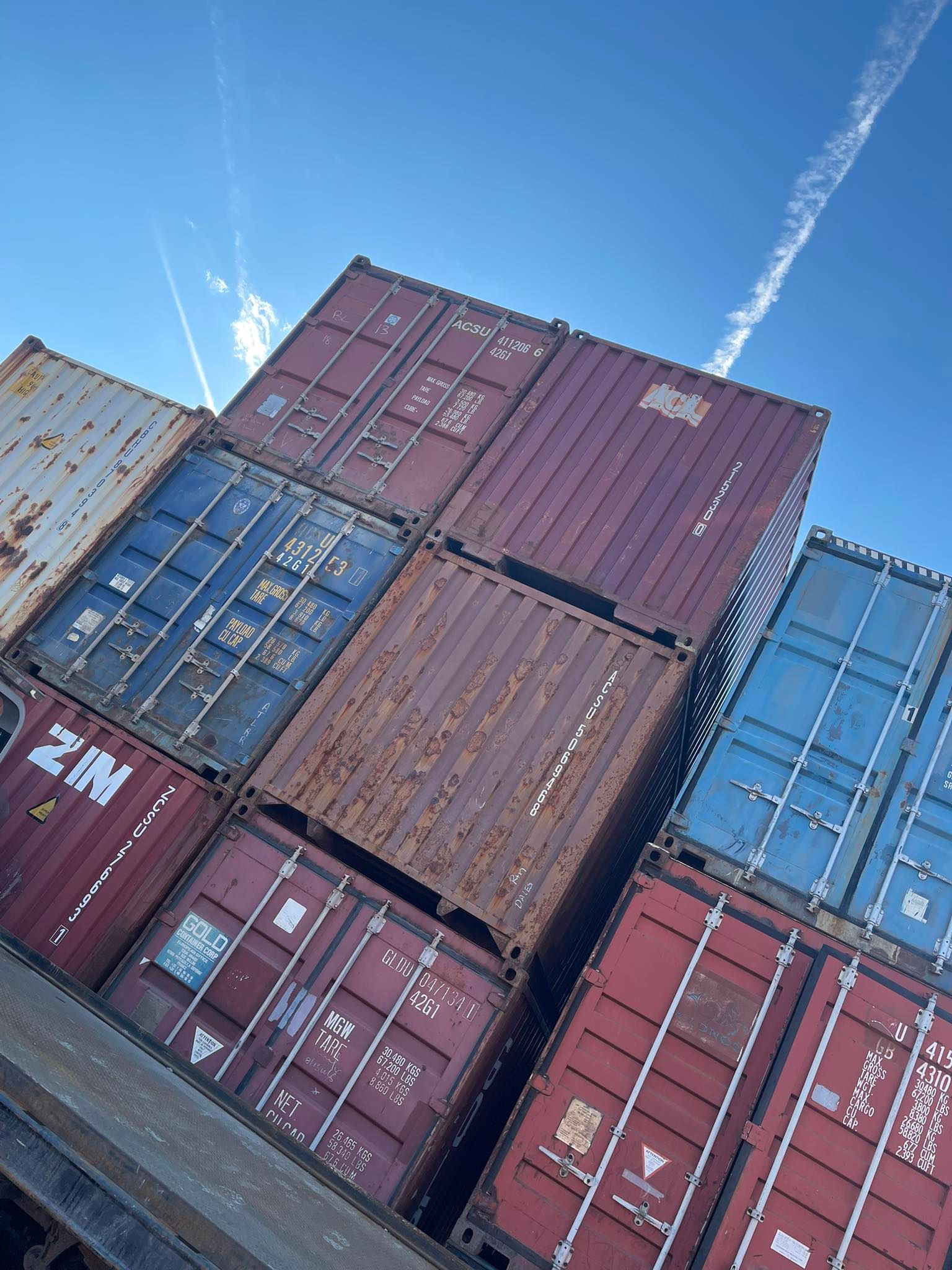 A low-angle view of stacked red, blue, and weathered shipping containers against a clear blue sky with a contrail.
